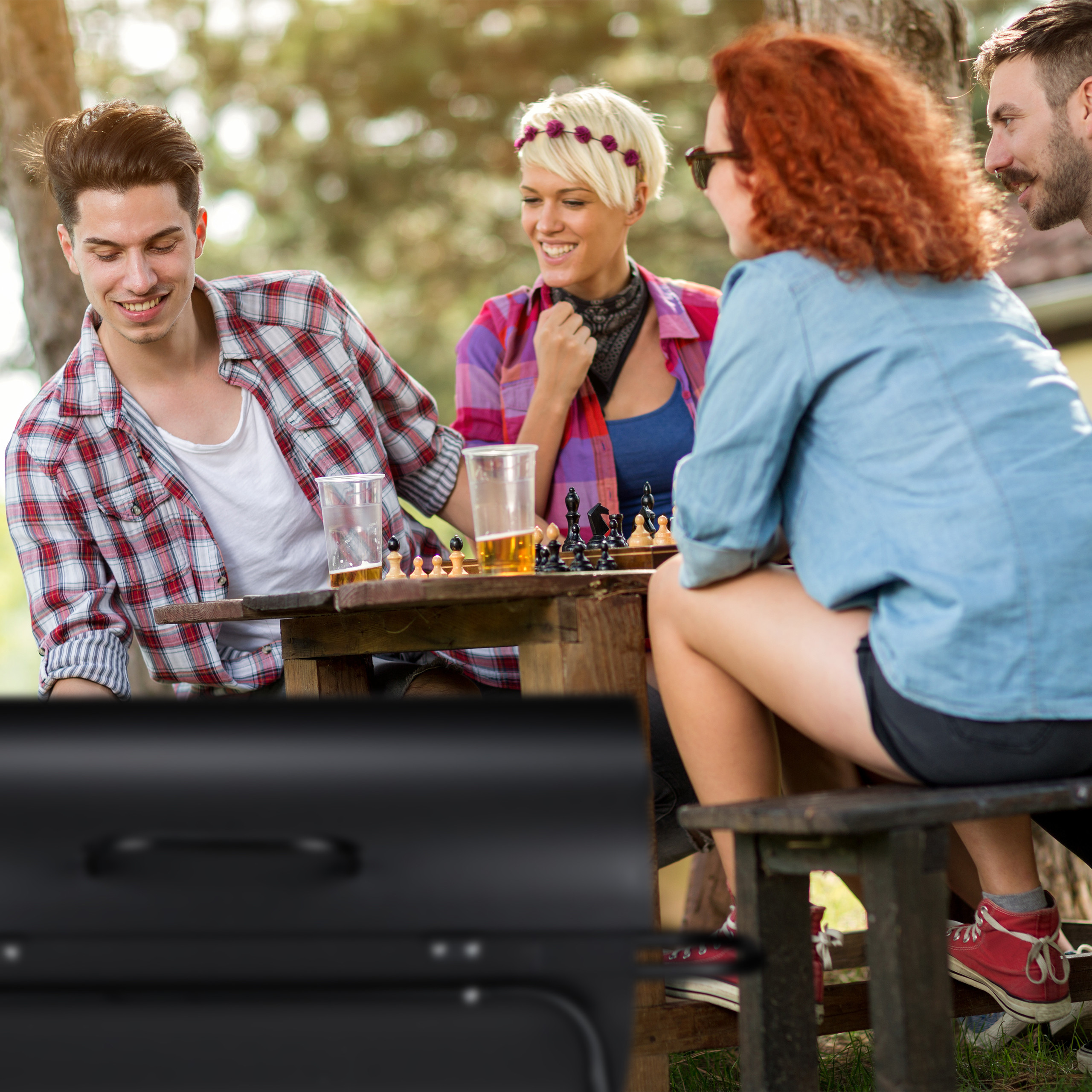 Vier Freunde spielen Schach an einem Holztisch im Freien, daneben steht ein Grill.