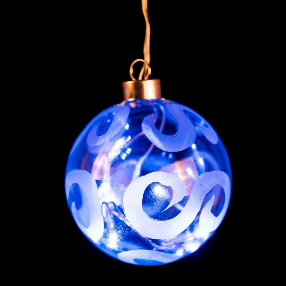 Un adorno navideño azul con patrones blancos y luces brillantes en su interior cuelga sobre fondo oscuro.