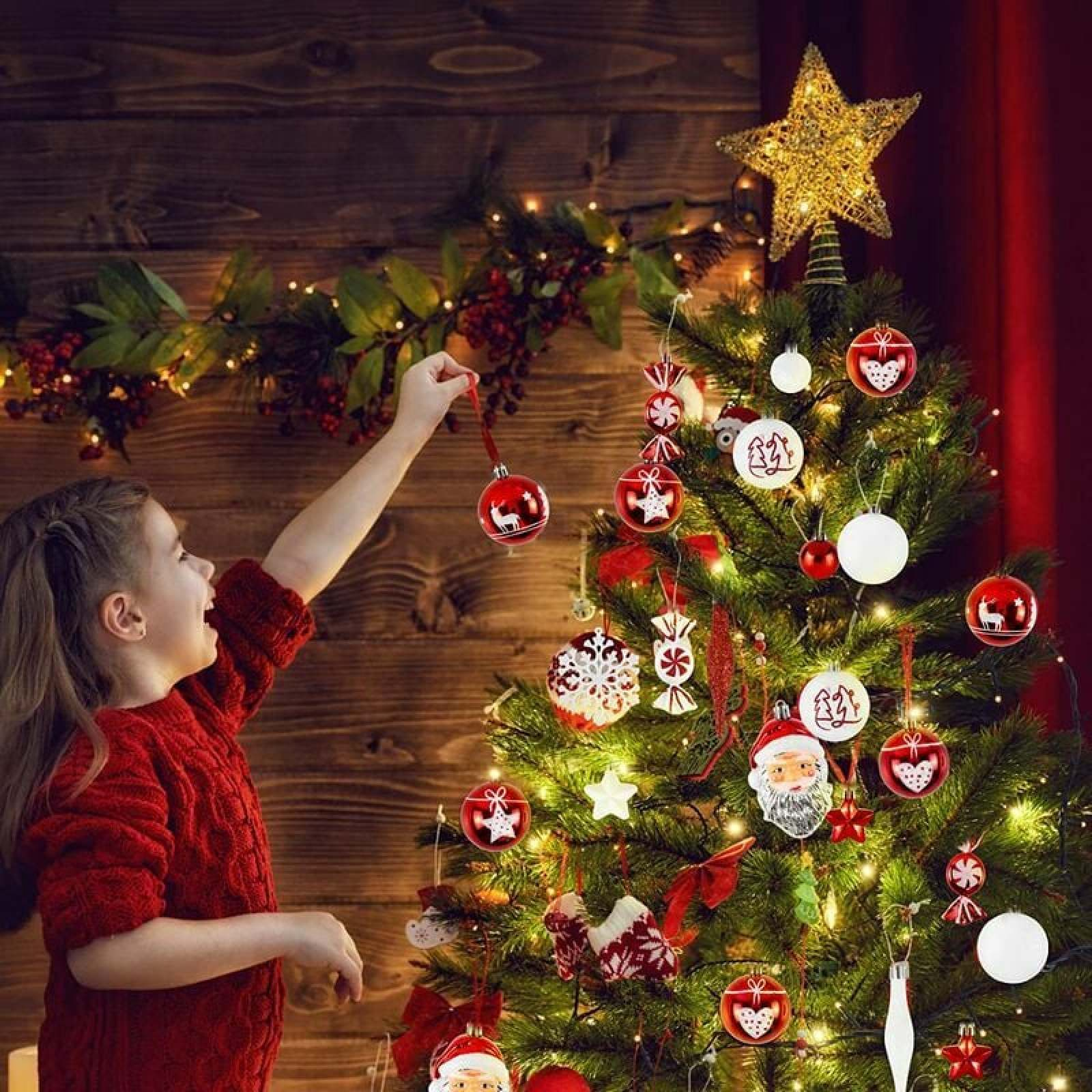 Ein junges Mädchen in einem roten Pullover hängt eine rote Kugel an einen geschmückten Weihnachtsbaum.