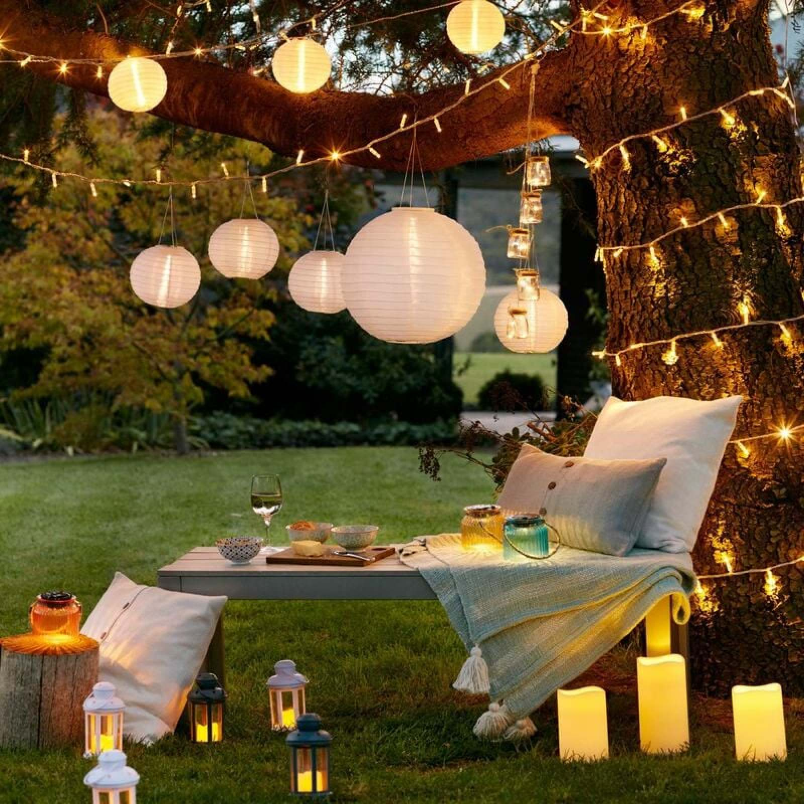 Beleuchteter Baum mit Lampions und Lichterketten über einer gemütlichen Bank im Garten.