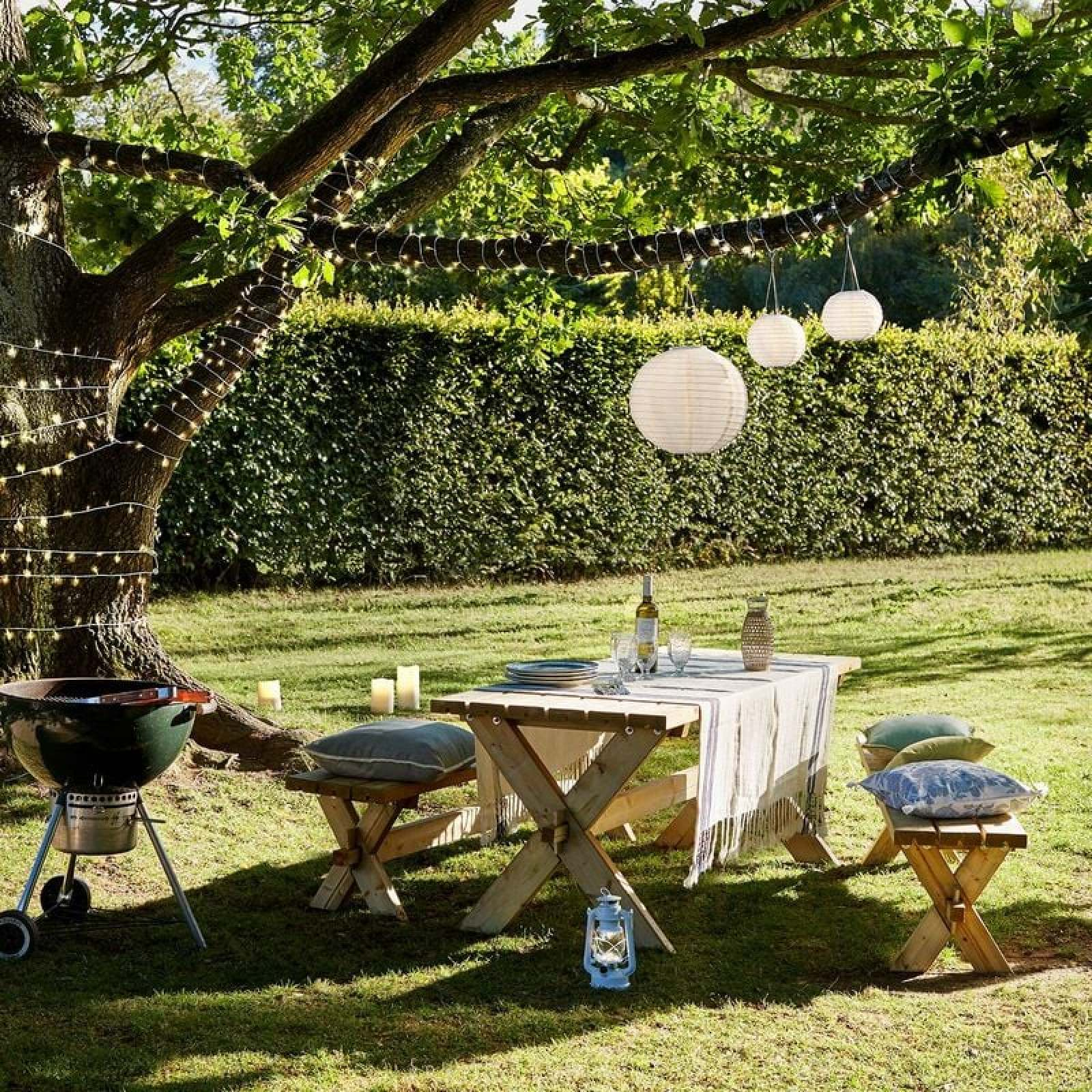 Ein hölzernen Tisch unter einem beleuchteten Baum, mit Laternen, Kissen und einem Grill im Garten.