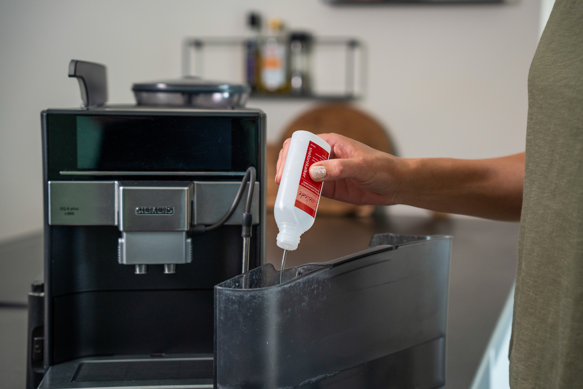 A person pours descaler from a white bottle into the water tank of a Siemens coffee machine.