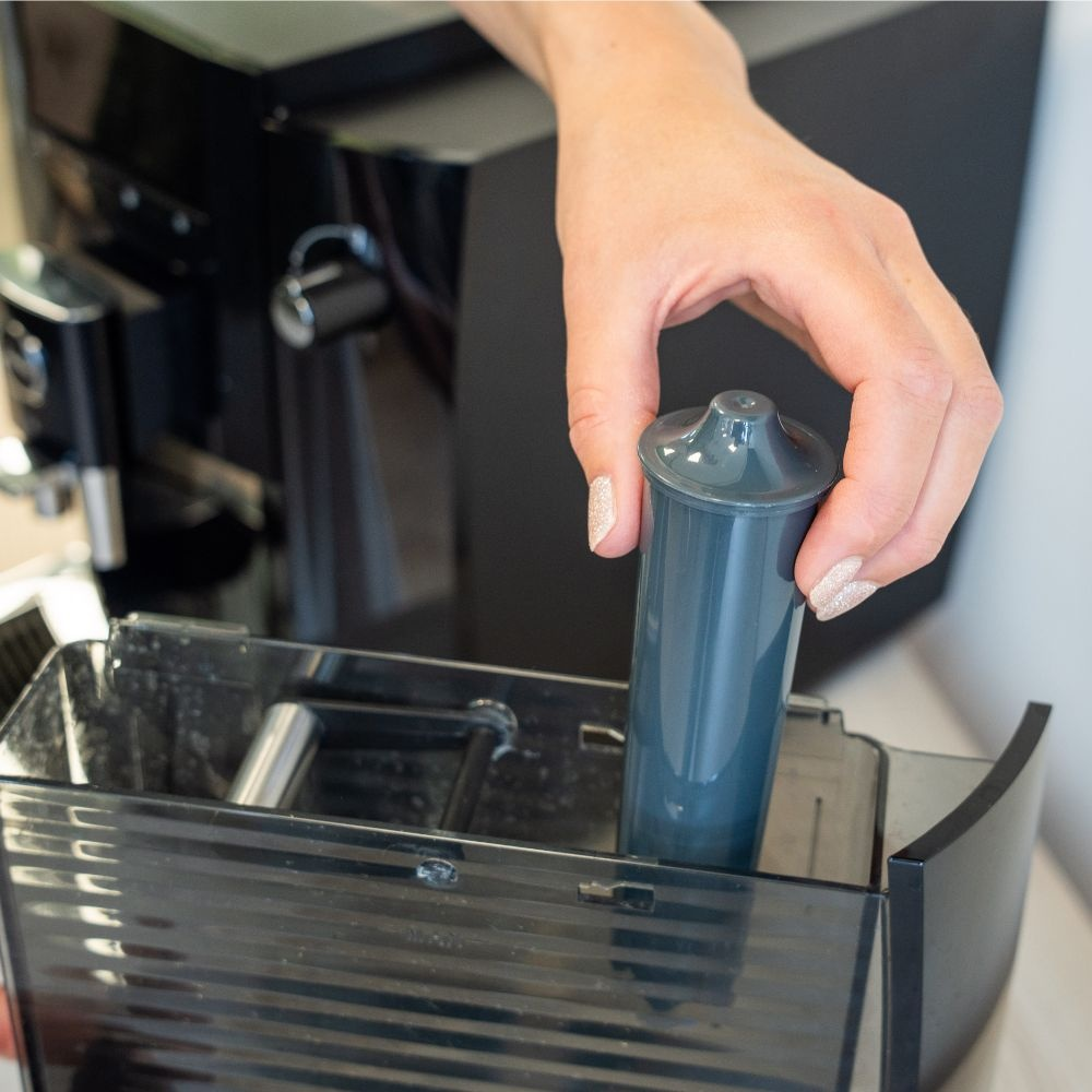 A hand inserts a grey water filter into a coffee machine water tank.