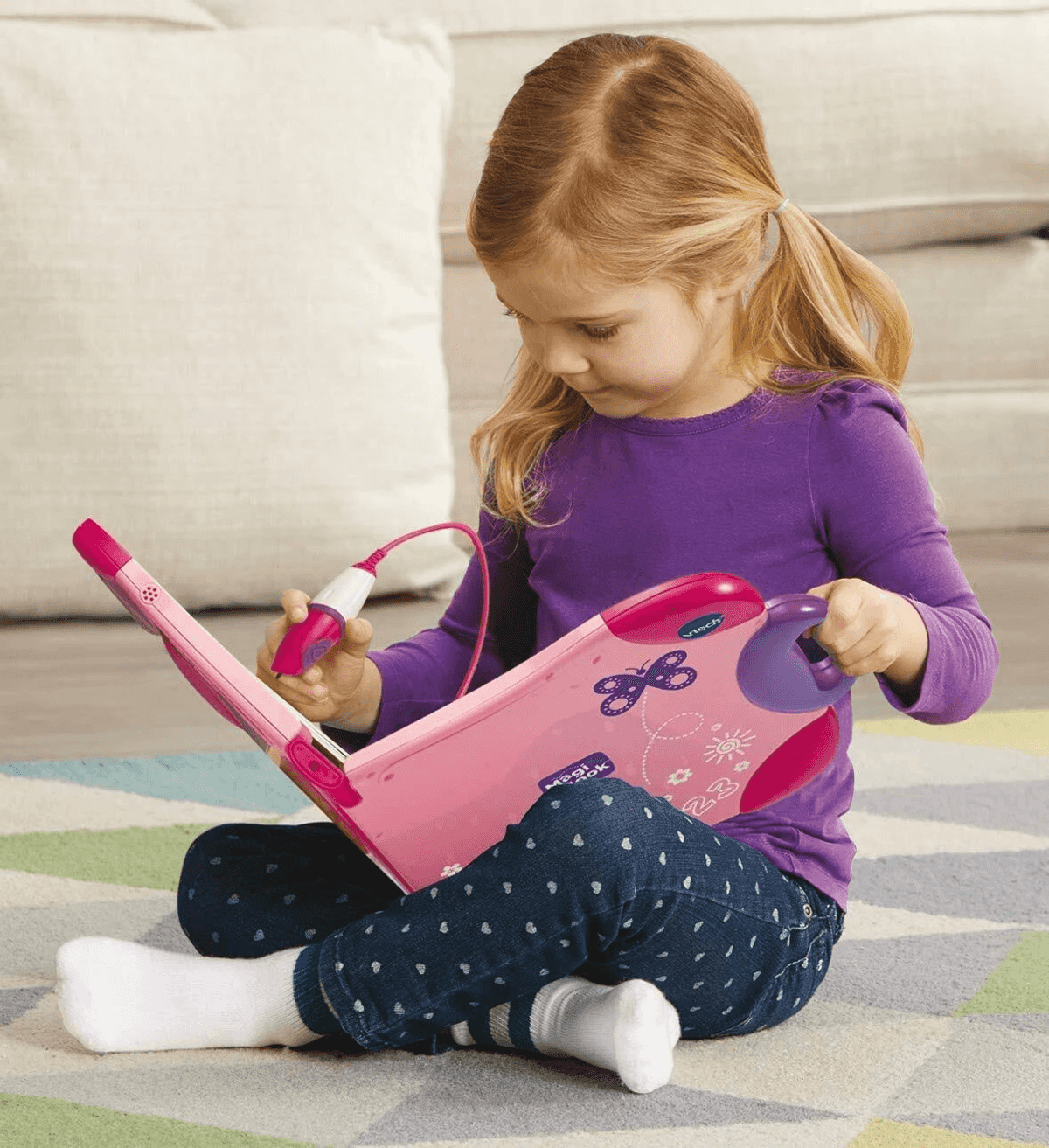 Una niña con una camisa morada se sienta en una alfombra con patrones, usando un libro de juguete interactivo rosa.