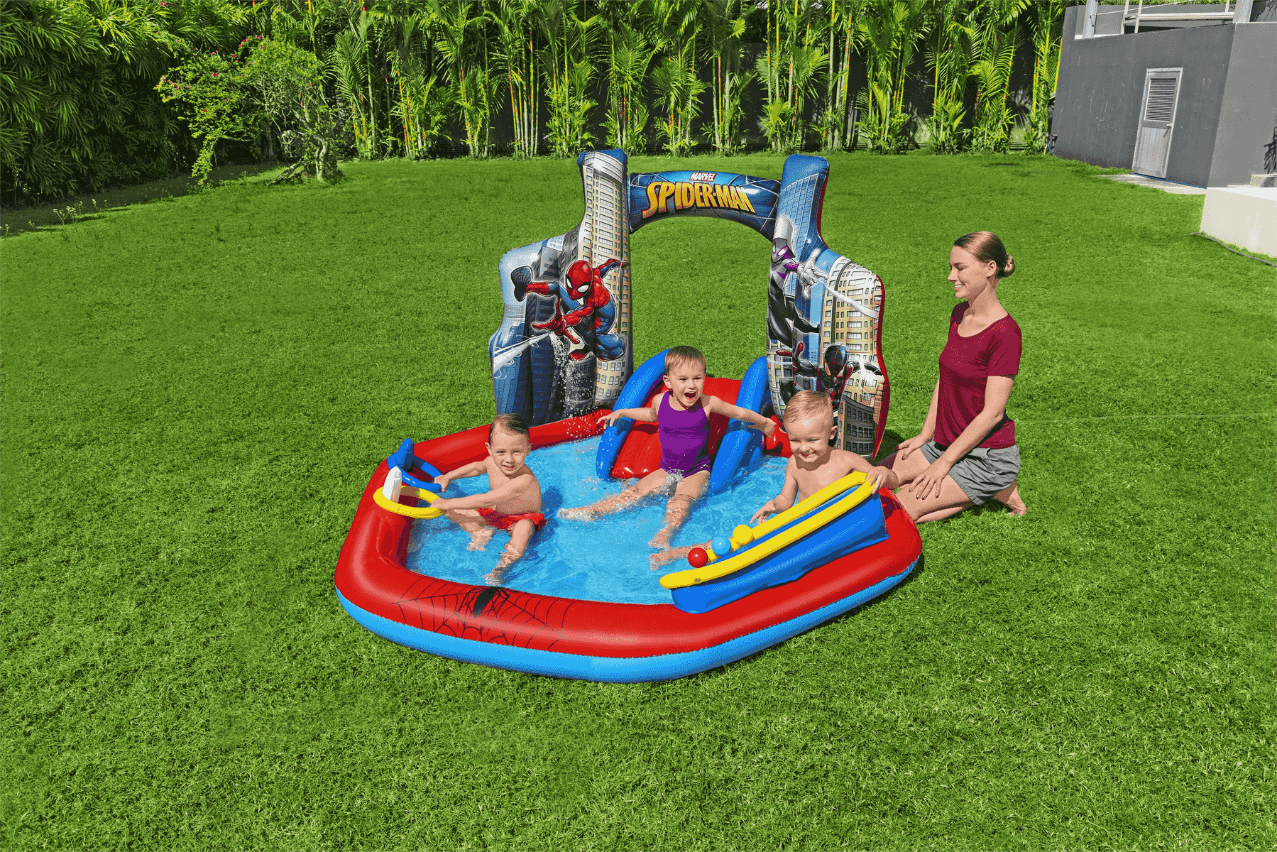 Tres niños juegan en una piscina inflable colorida de Spider-Man en el césped mientras una mujer observa.
