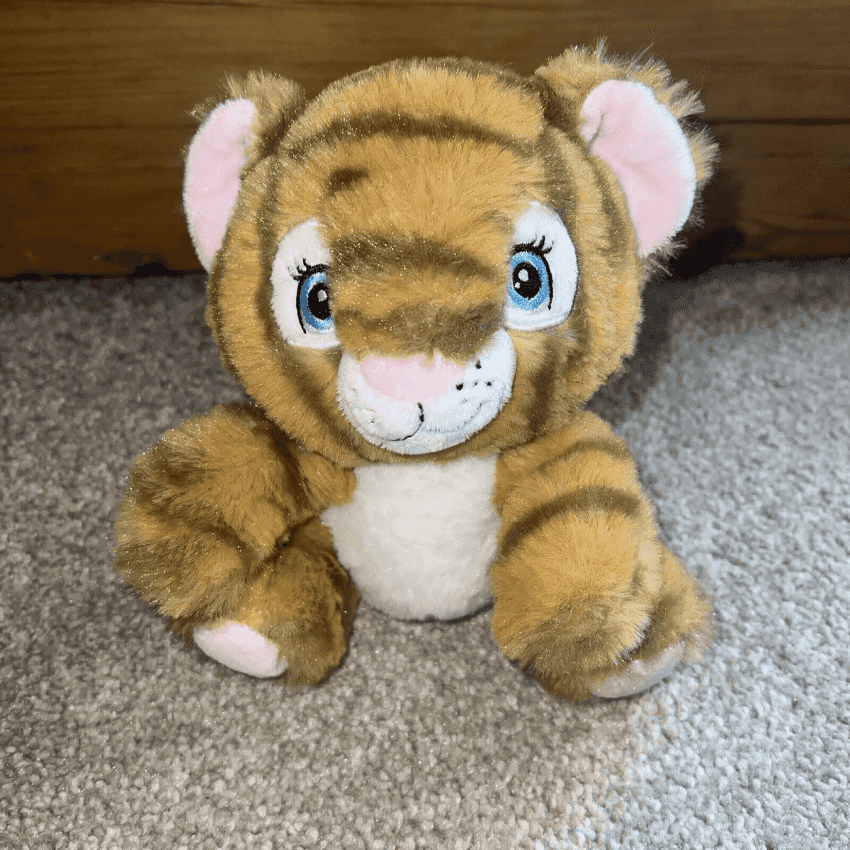 Un tigre de peluche con ojos azules y rayas marrones se sienta en una alfombra gris.