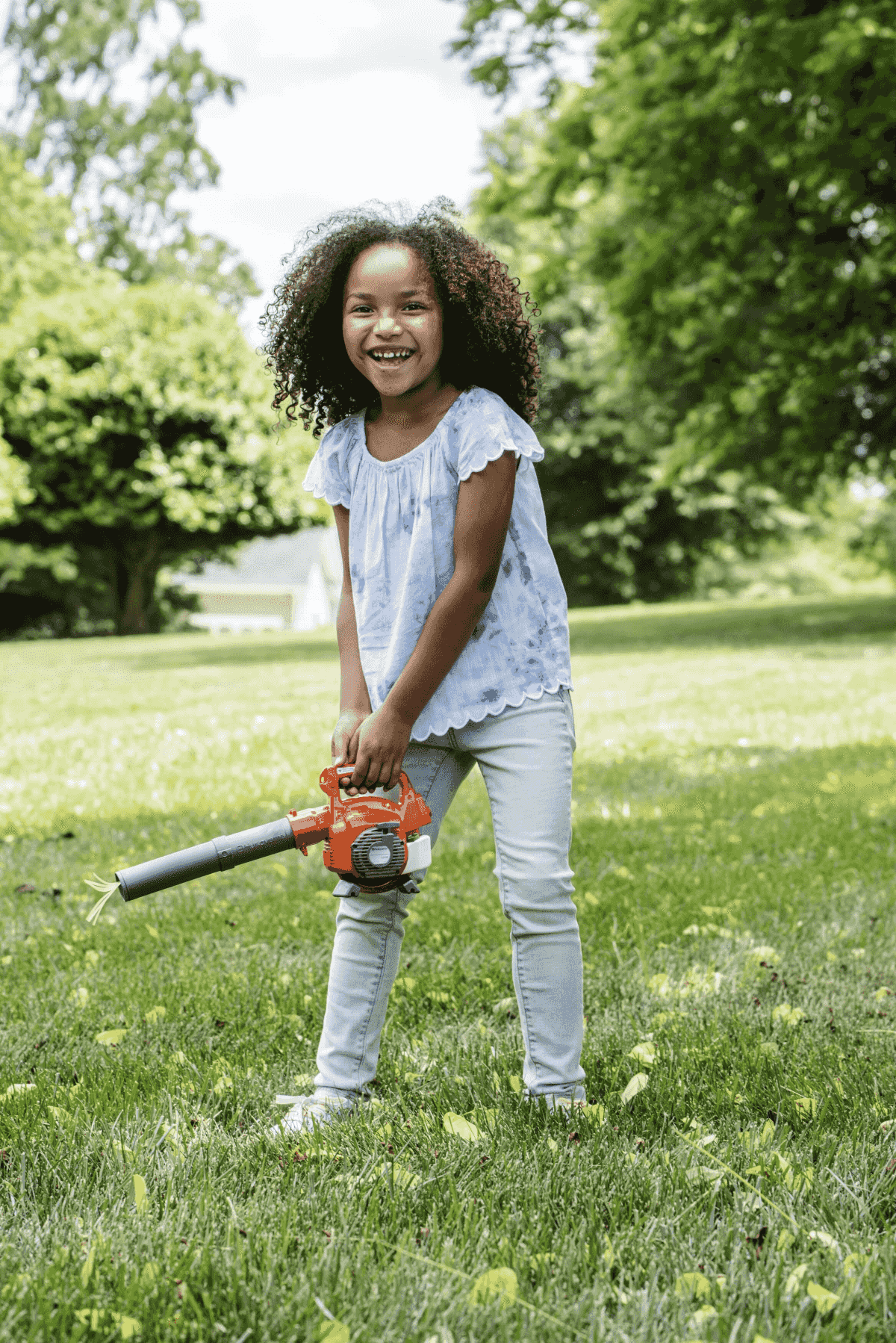 Una niña sonriente con cabello rizado sostiene un soplador de hojas de juguete en un jardín verde.