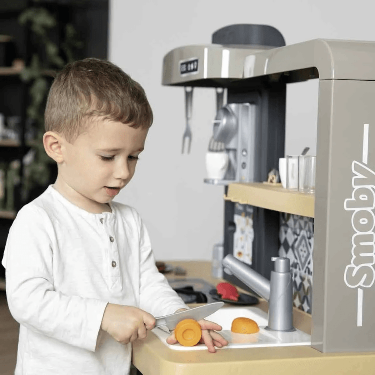 Un niño pequeño con camisa blanca corta una zanahoria de juguete en una cocina de juguete.