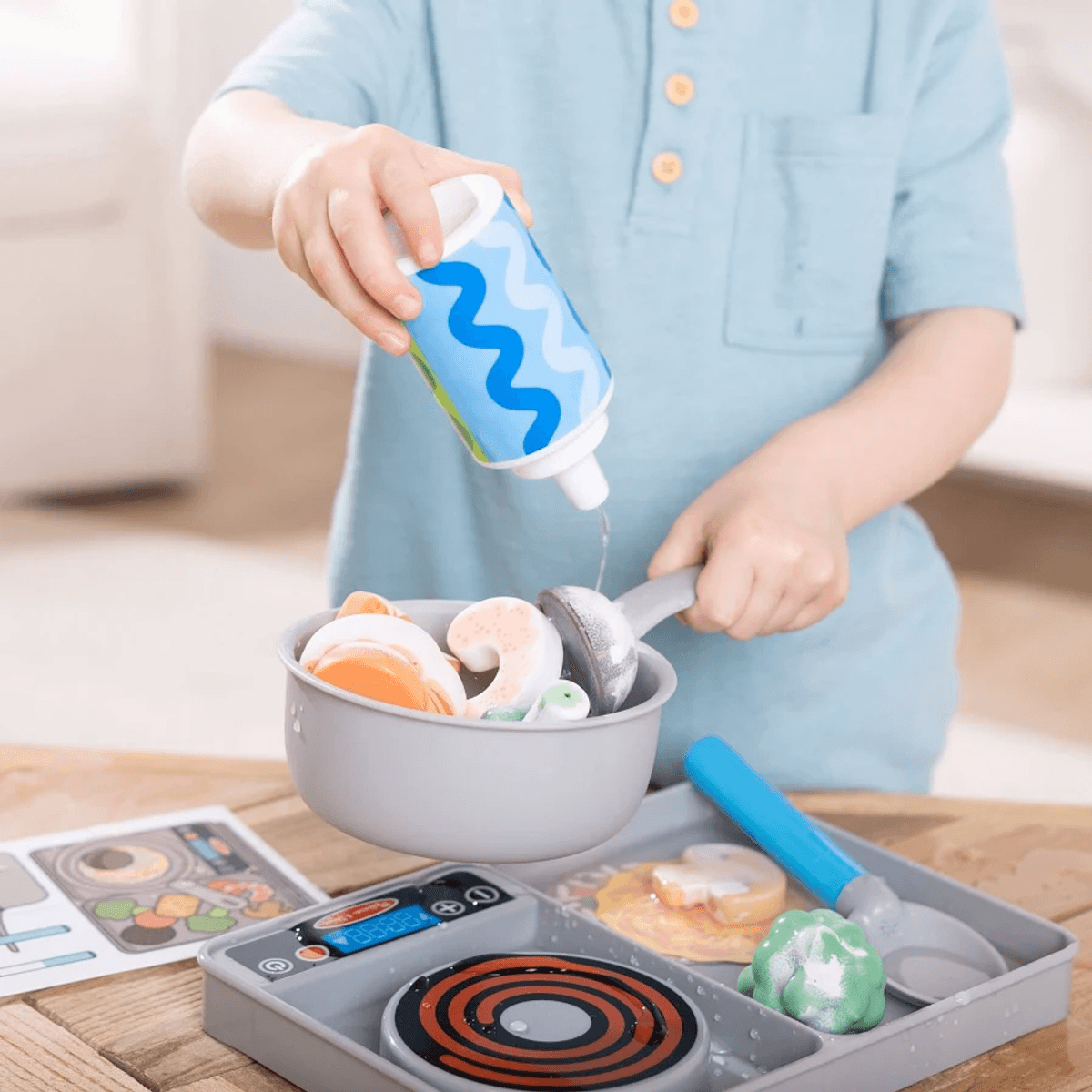 Un niño con camisa azul vierte agua de una botella de juguete en una olla gris con comida de juguete.