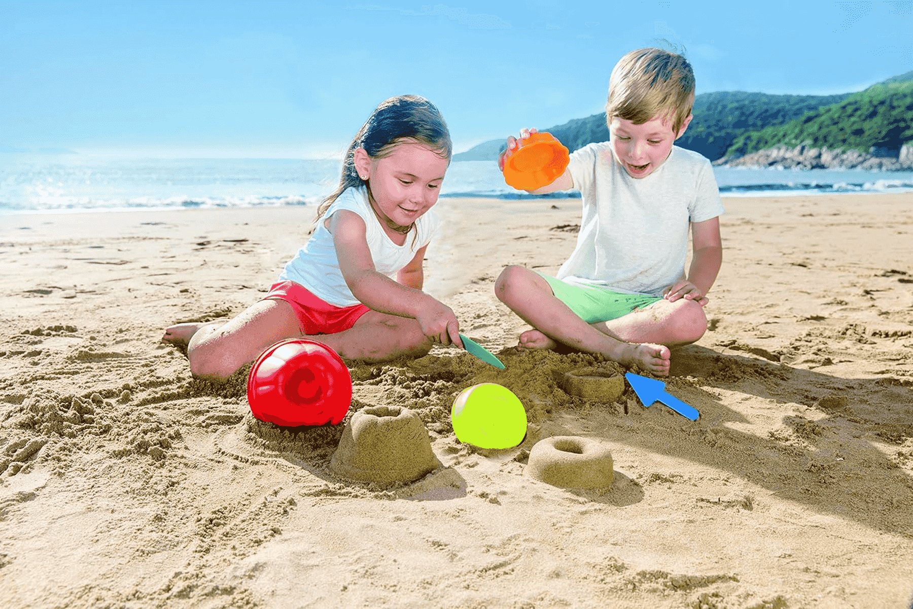 Dos niños jugando con juguetes de arena en una playa soleada, con el mar y colinas al fondo.