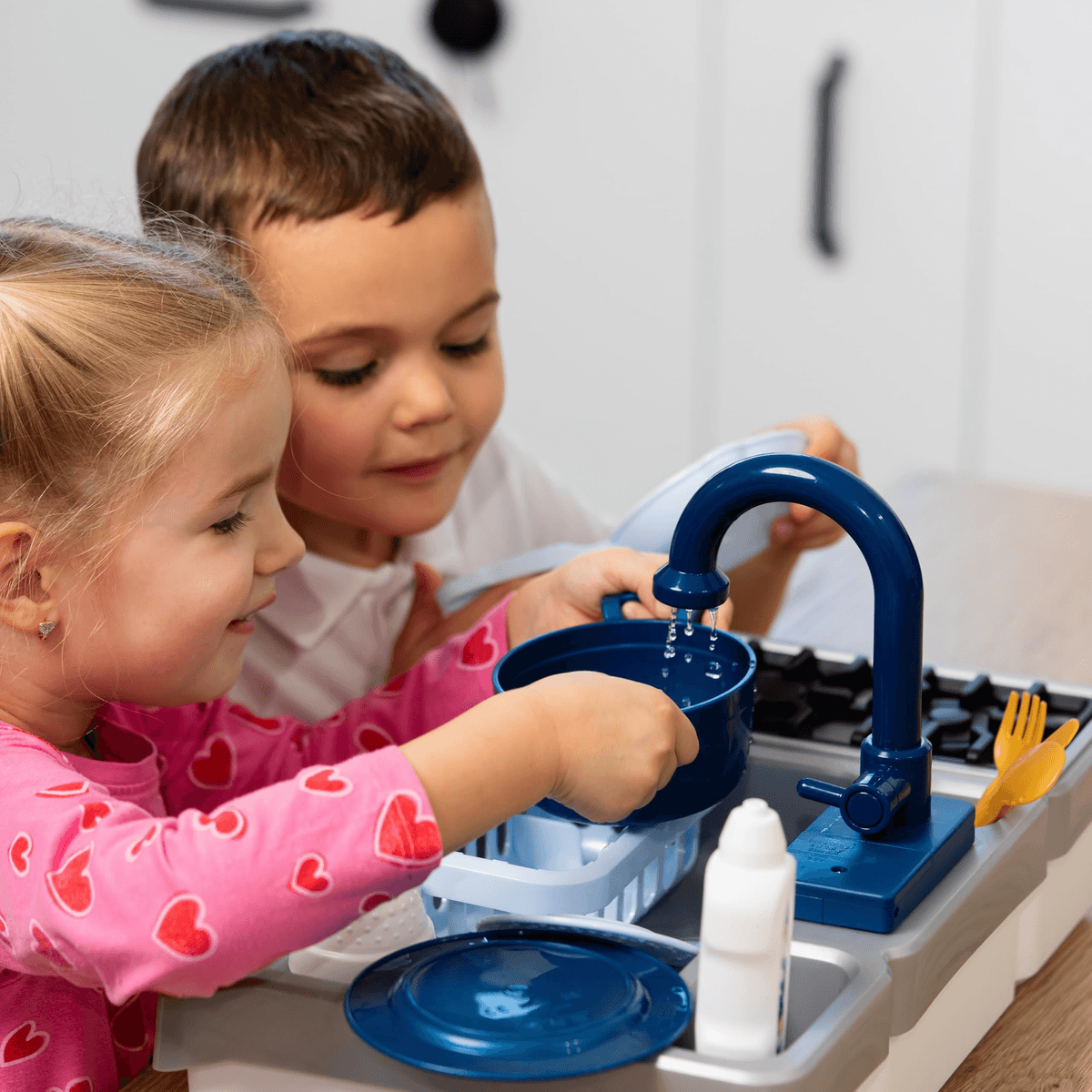 Los niños juegan con un fregadero de juguete azul, lavando platos en una cocina luminosa.
