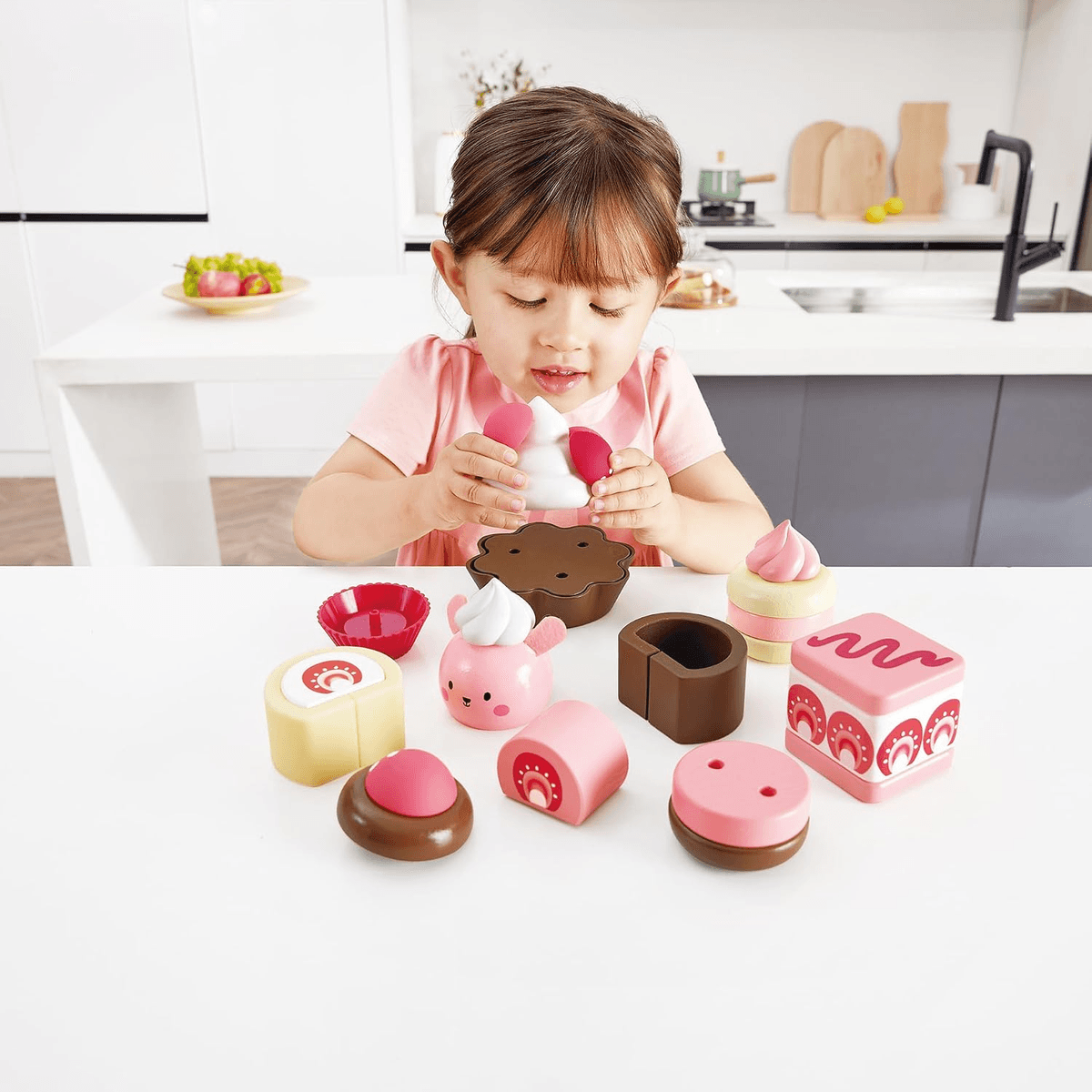 Una niña con camisa rosa juega con postres de juguete de madera en una mesa blanca.