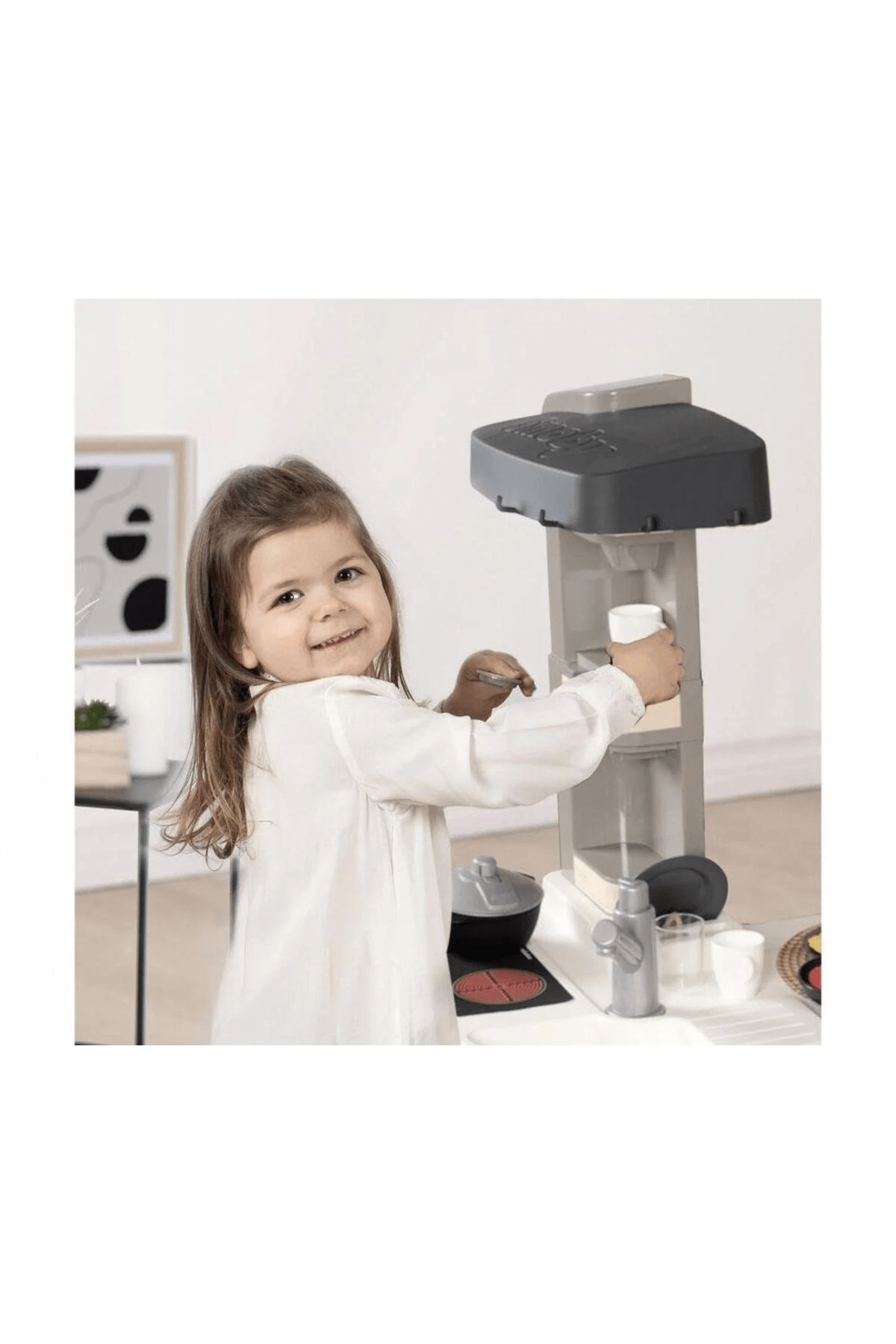 Una niña pequeña con blusa blanca juega sonriente con una cocinita de juguete gris.