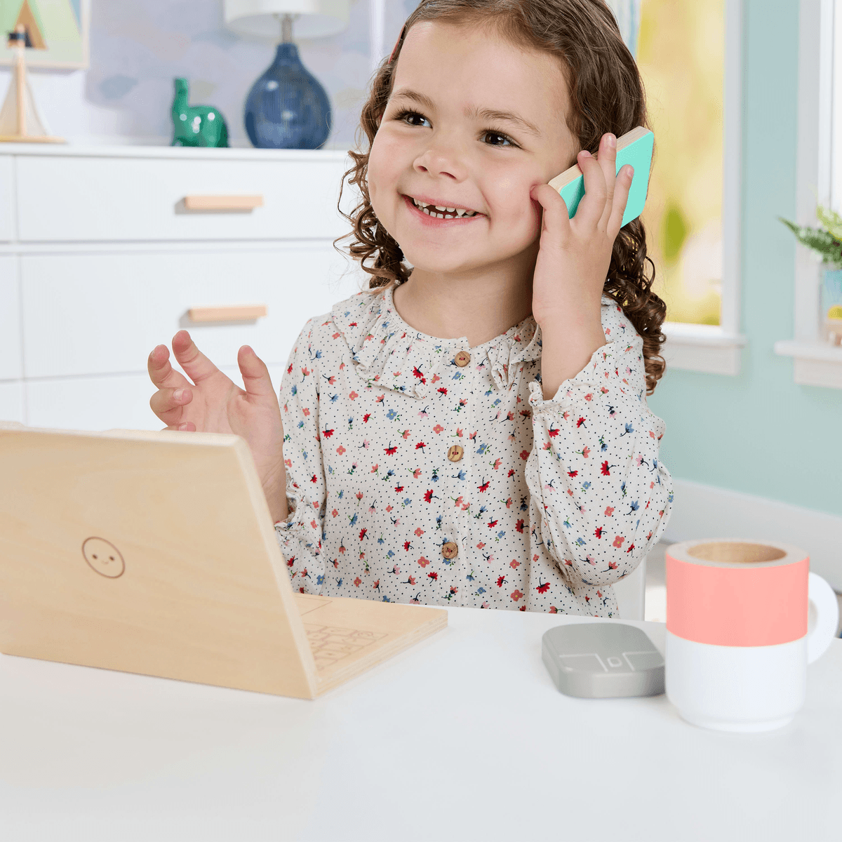 Niña sonriente con blusa floral hace llamada con juguete de madera en mesa.