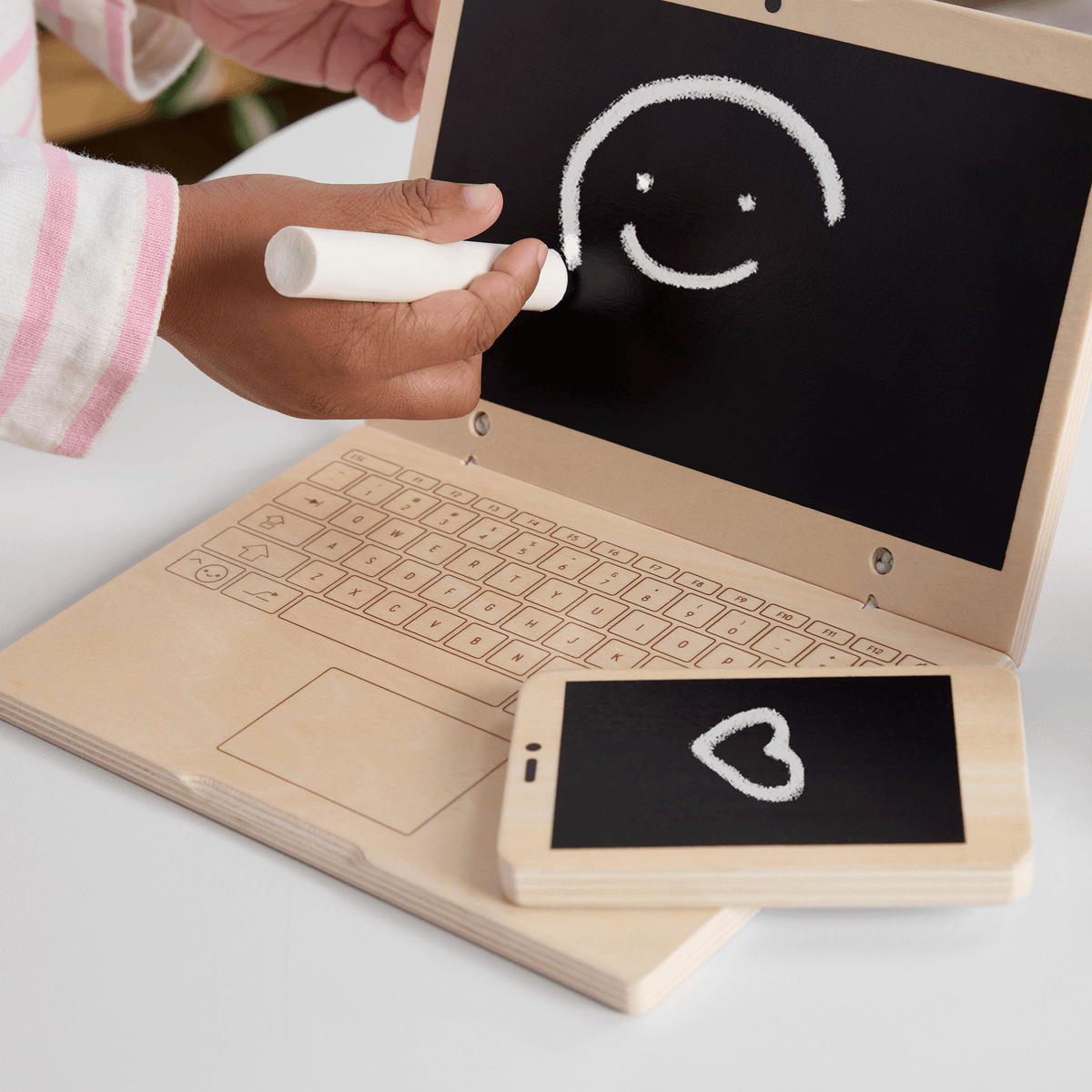 Un niño dibuja una carita feliz con tiza en una laptop de juguete de madera junto a un teléfono de juguete.