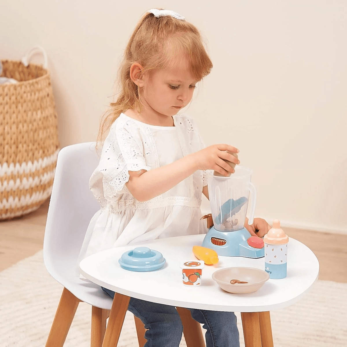 Una niña pequeña está sentada en una mesa blanca, jugando con una licuadora de juguete azul y accesorios de madera.