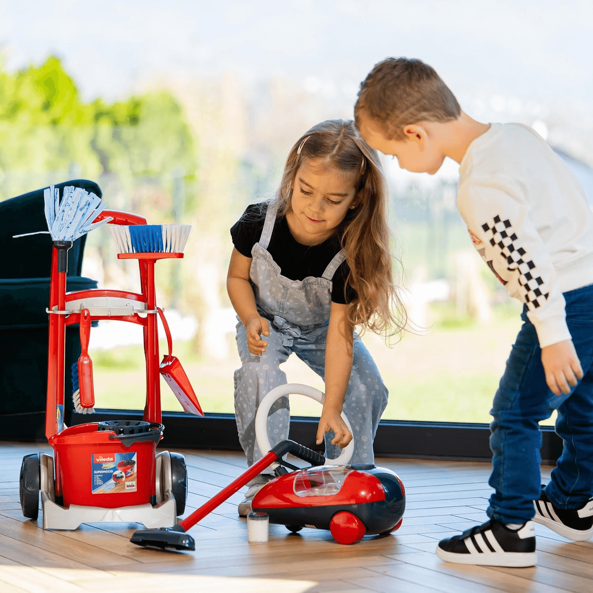 Zwei Kinder spielen drinnen mit einem roten Spielzeugstaubsauger und Reinigungswagen.
