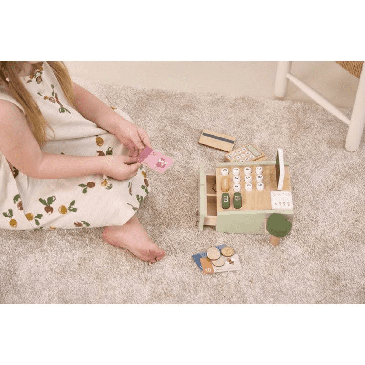 Una niña jugando en una alfombra beige con una caja registradora de juguete de madera, dinero y tarjetas.