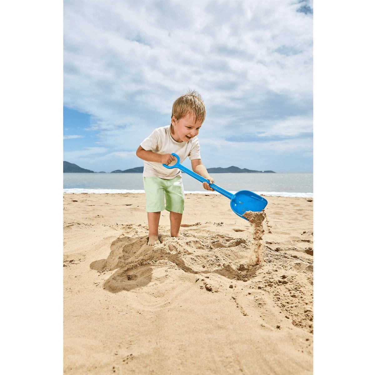 Un niño sonriendo mientras cava arena con una pala azul en la playa.
