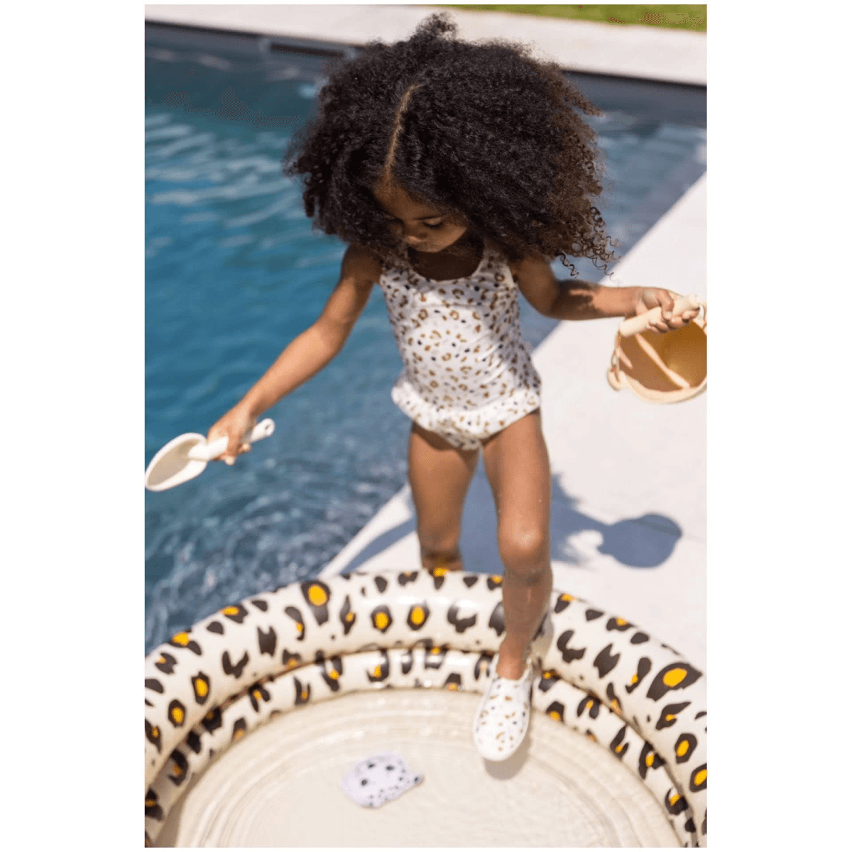 Una niña pequeña con cabello oscuro rizado está en una piscina infantil con estampado de leopardo.