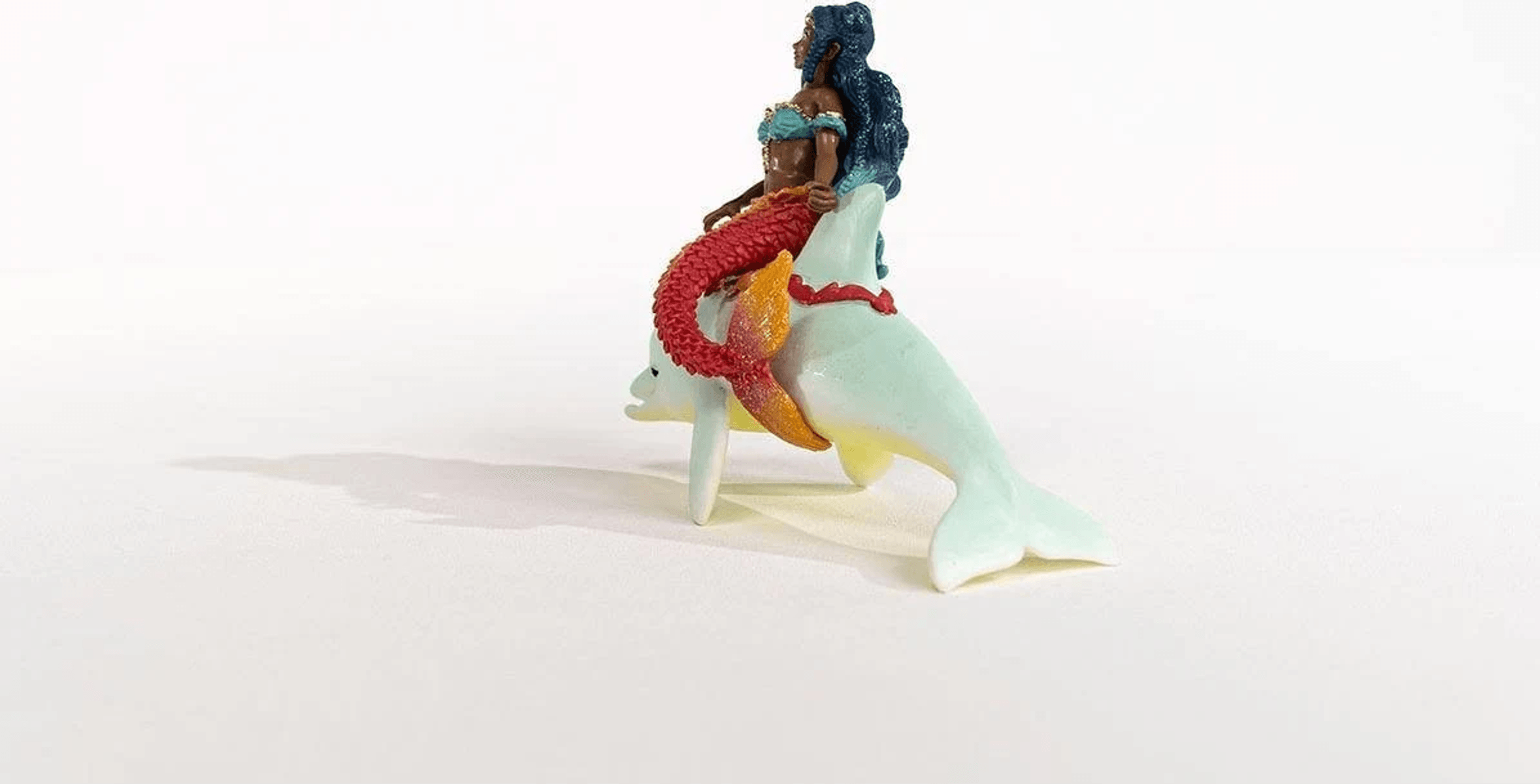 Una pequeña figura muestra a una sirena con cola roja y pelo largo montada en un delfín blanco sobre fondo blanco.
