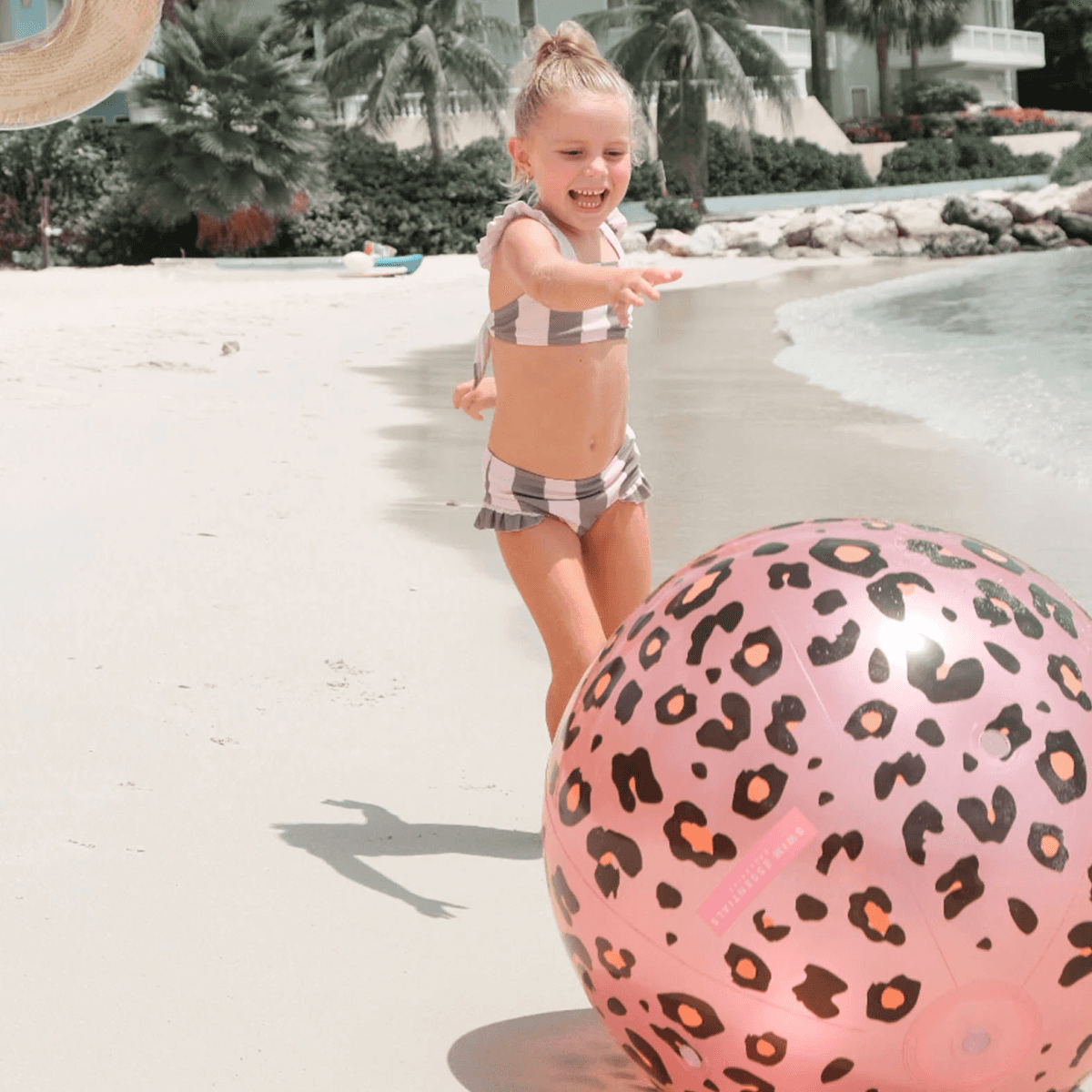 Una niña en bikini de rayas juega con una gran pelota de playa con estampado de leopardo rosa en una playa soleada.