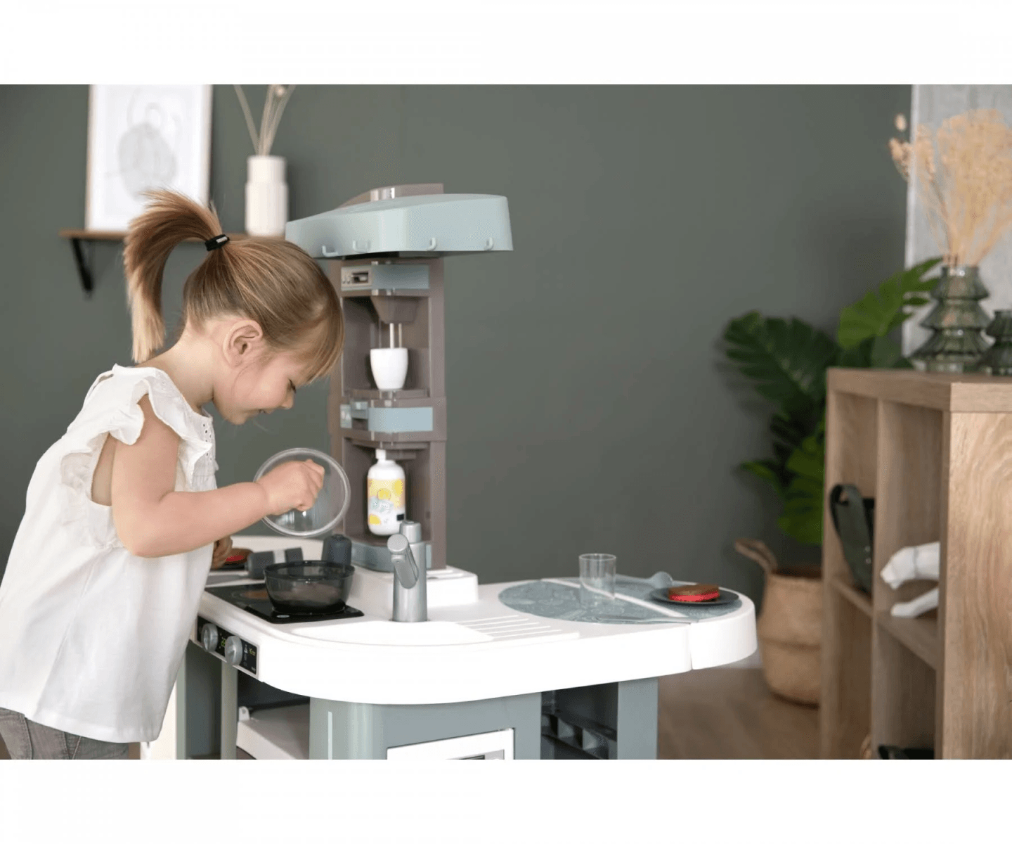 Una niña con una blusa blanca juega con una cocinita de juguete en una habitación con paredes verdes.