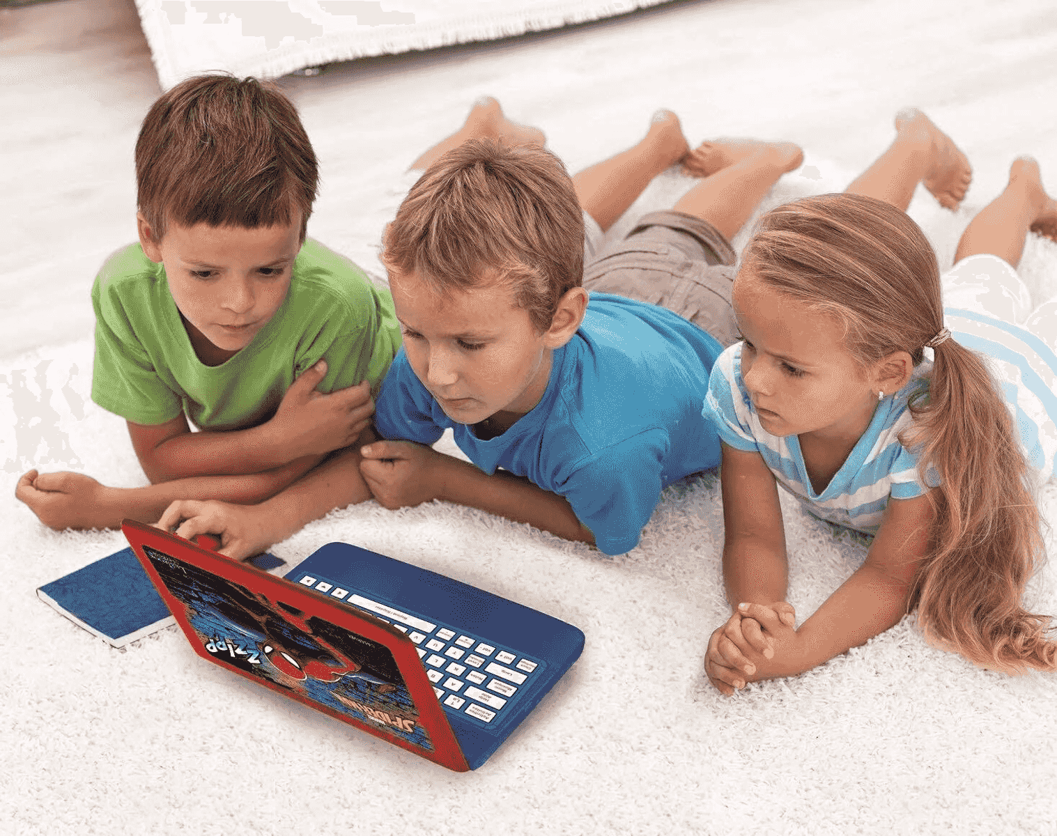 Tres niños yacen sobre una alfombra blanca, mirando fijamente una colorida computadora portátil de juguete.
