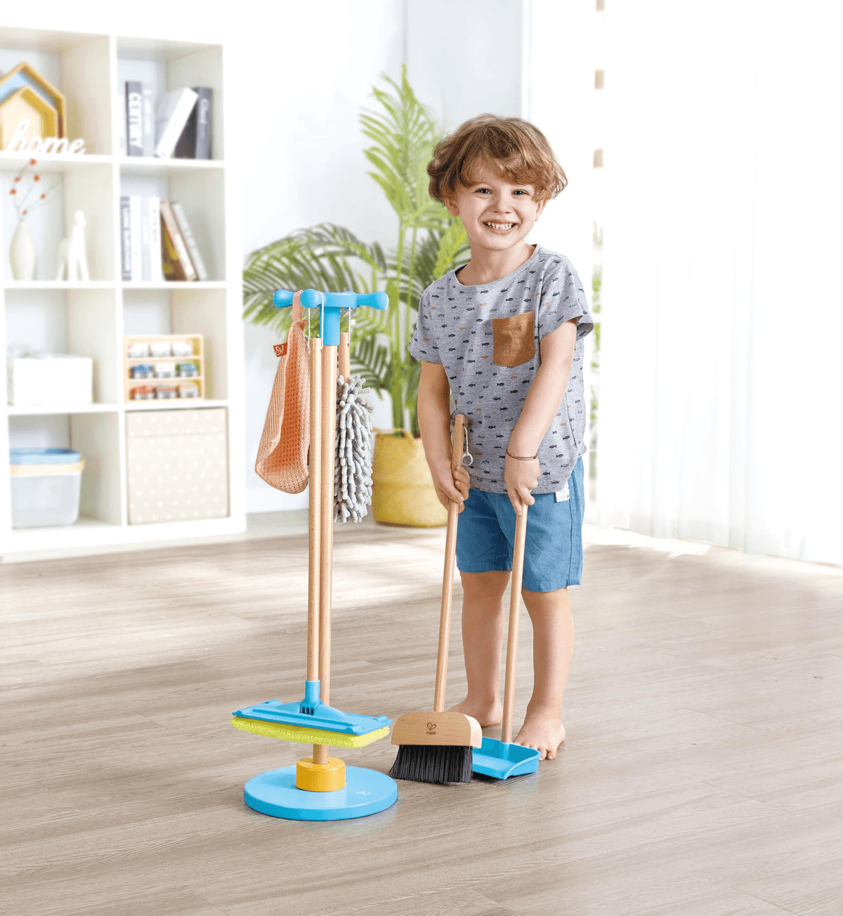 Un niño sonriente está en una habitación sosteniendo una escoba y un recogedor junto a un carrito de limpieza de juguete.