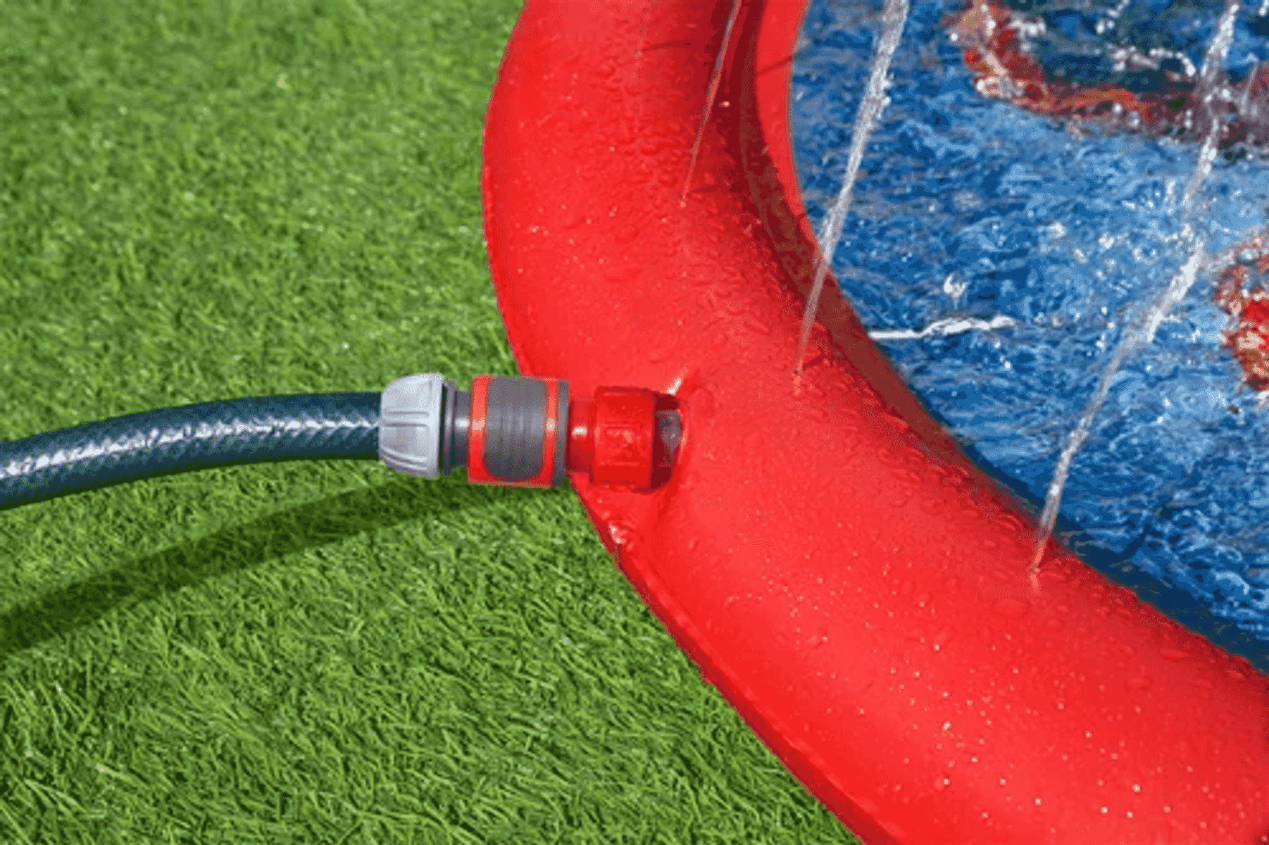 Una manguera de jardín está conectada a una piscina inflable roja con chorros de agua sobre césped verde.