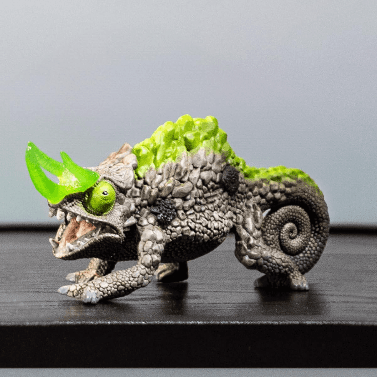 Una figura de lagartija de roca gris con cuernos, ojos y espinas de color verde brillante sobre una superficie oscura.