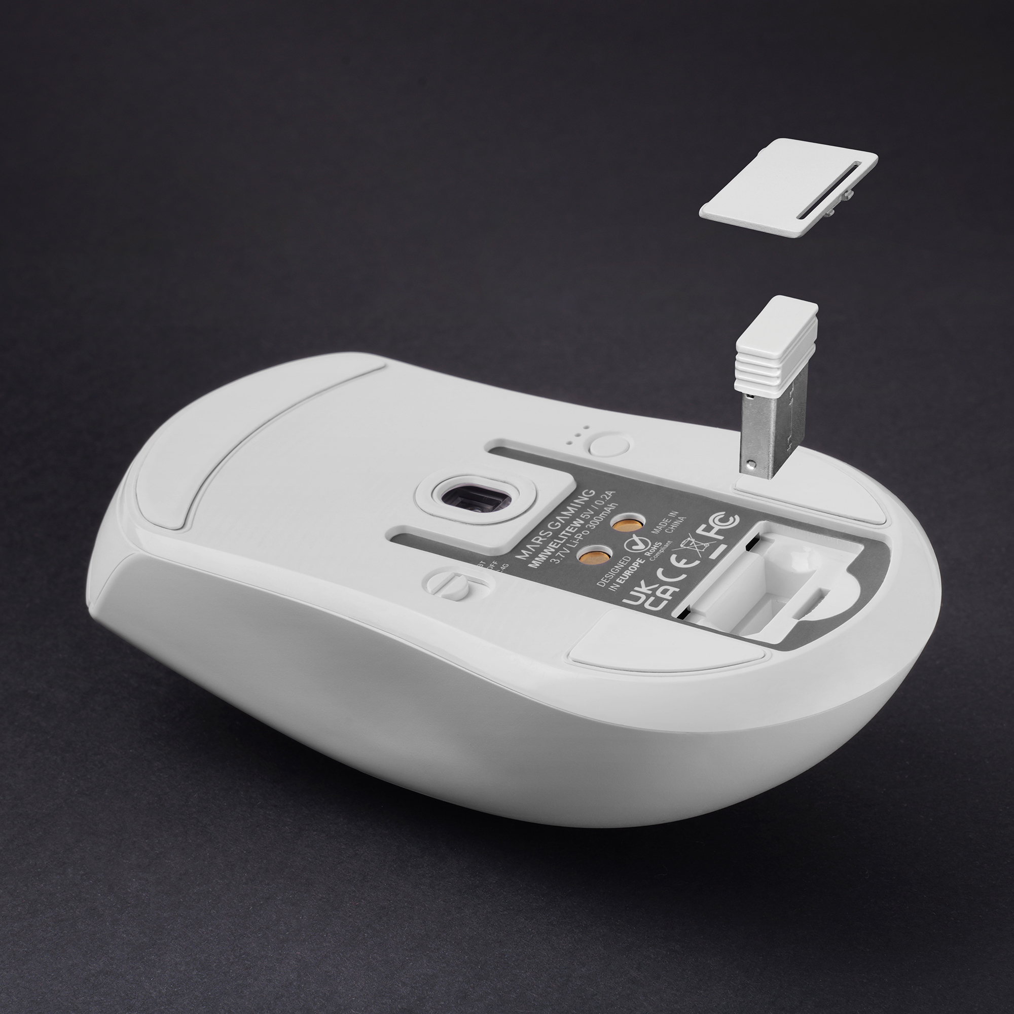 Parte inferior de un ratón blanco con el receptor USB y la tapa levantados sobre fondo oscuro.