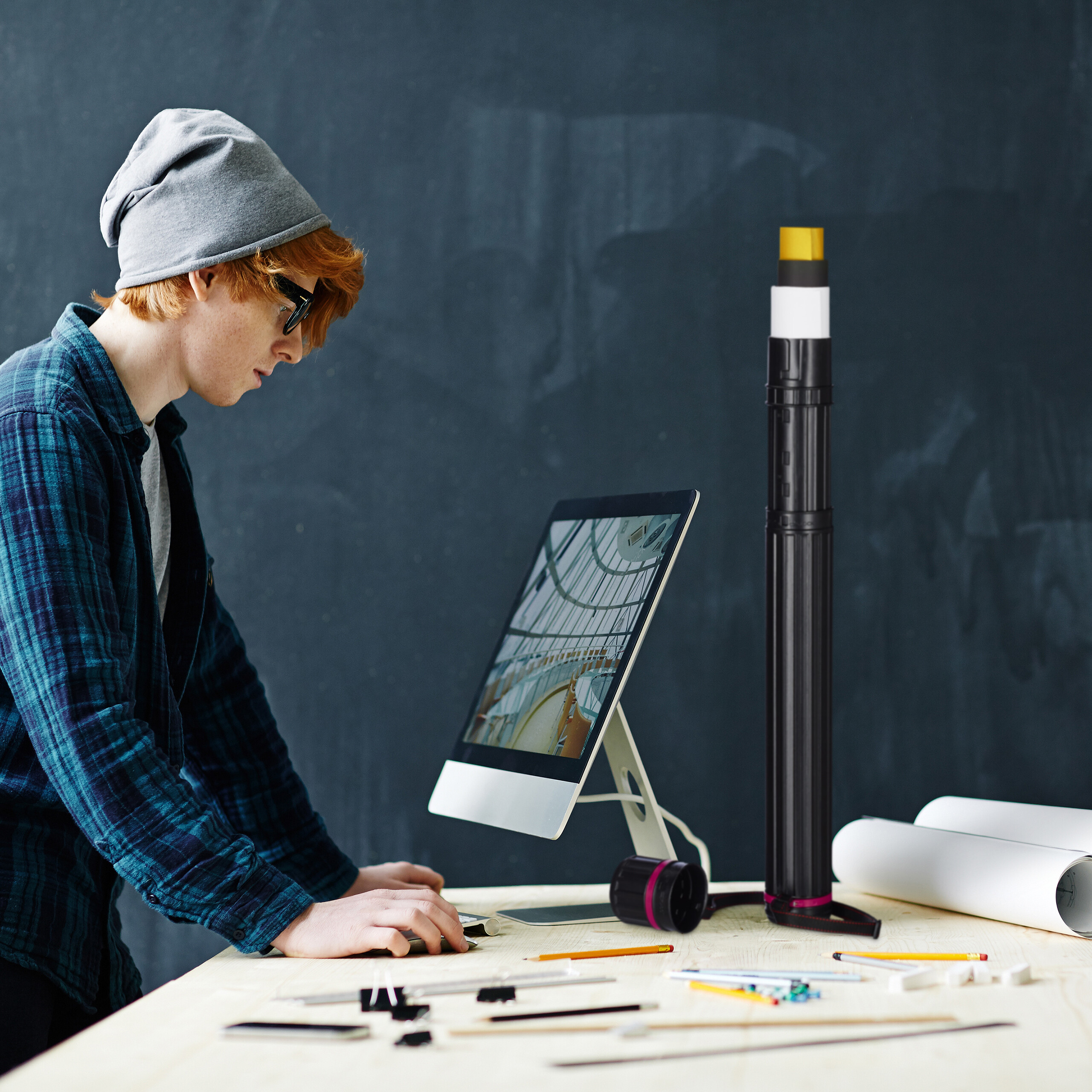 Ein junger Mann mit Mütze und kariertem Hemd arbeitet an einem Schreibtisch mit Architekturwerkzeugen.