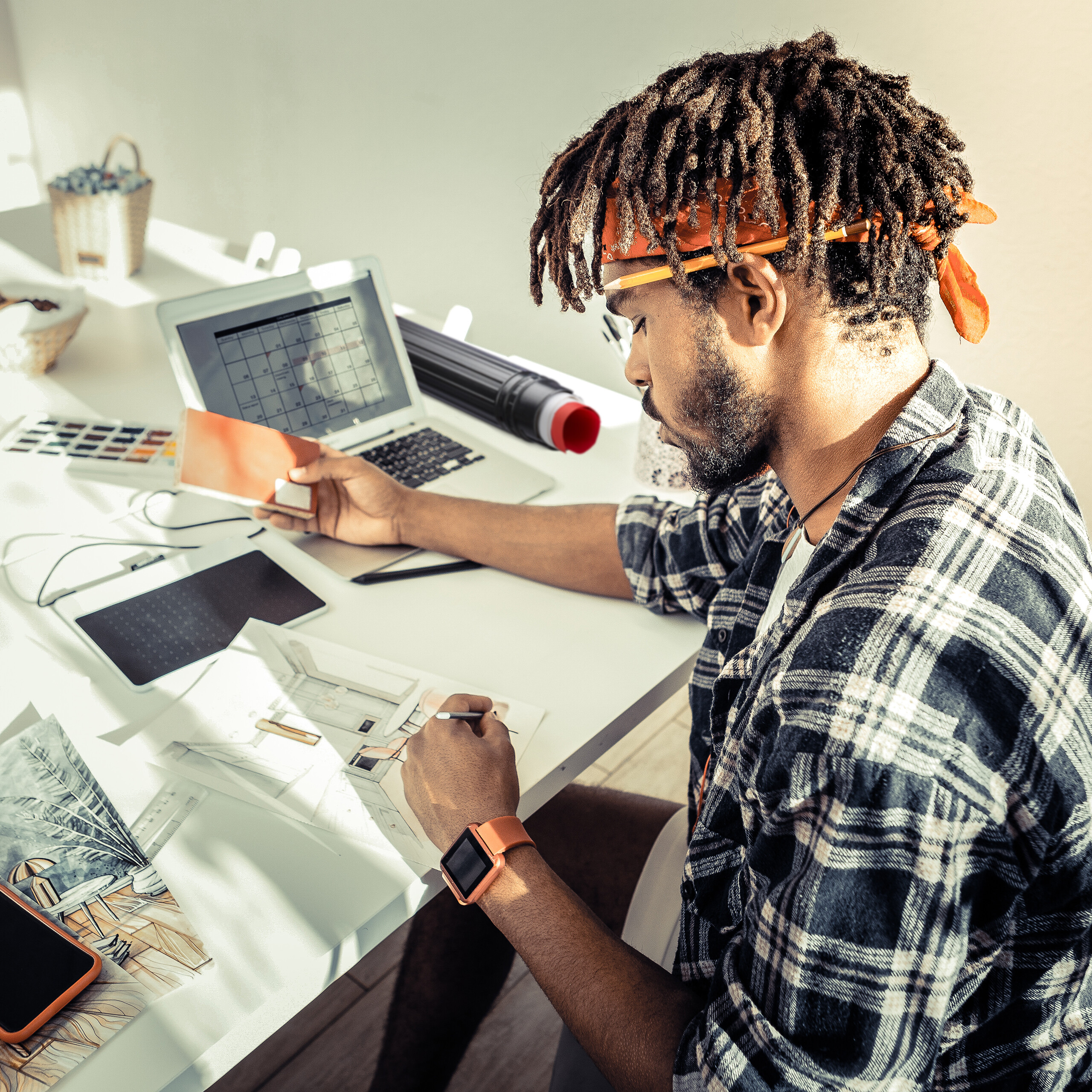 Ein Mann mit Dreadlocks und orangefarbenem Stirnband arbeitet an einem weißen Schreibtisch mit Laptop und Skizzen.