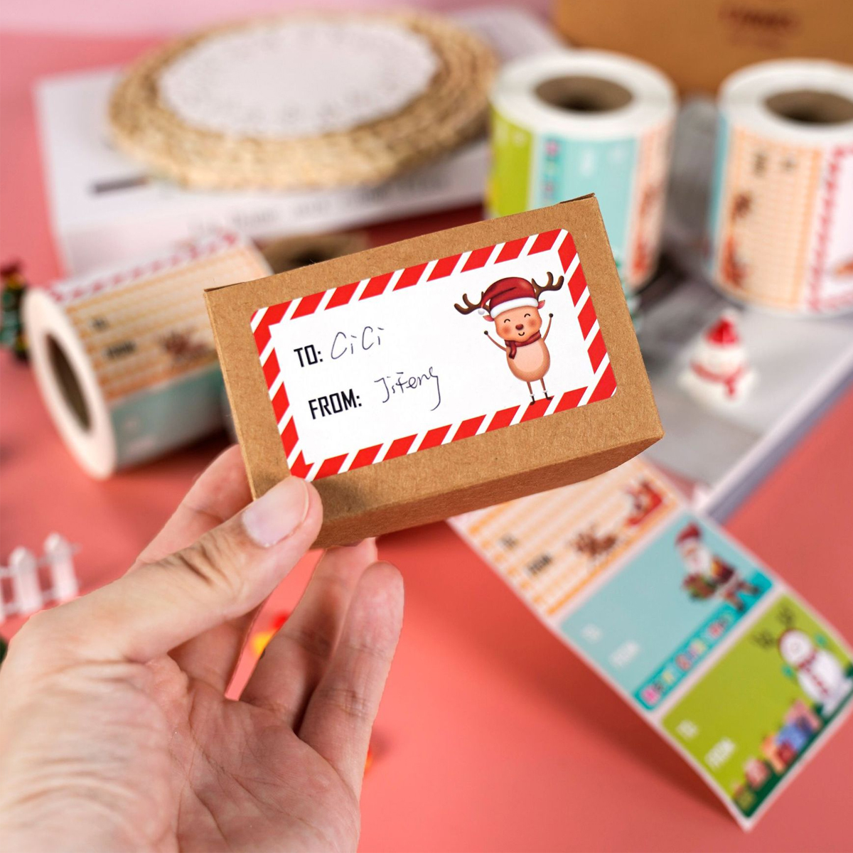 Hand hält kleines braunes Paket mit Weihnachts-Aufkleber, im Hintergrund Geschenkpapierrollen und Deko auf rosa Tisch.
