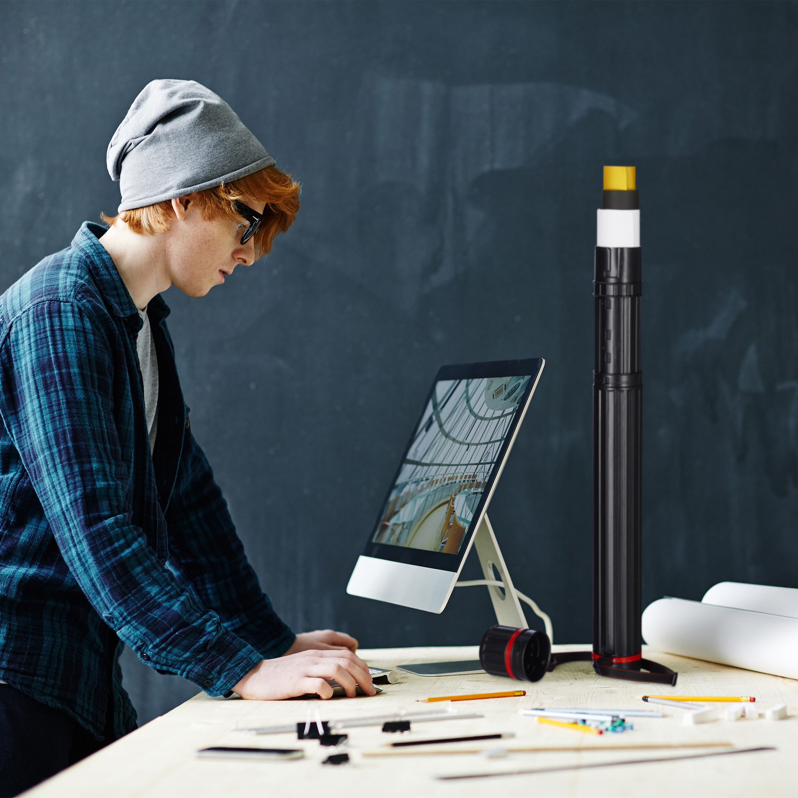Ein junger Designer mit Mütze arbeitet an einem Schreibtisch mit Computer, Blaupausen und Zeichenwerkzeugen.