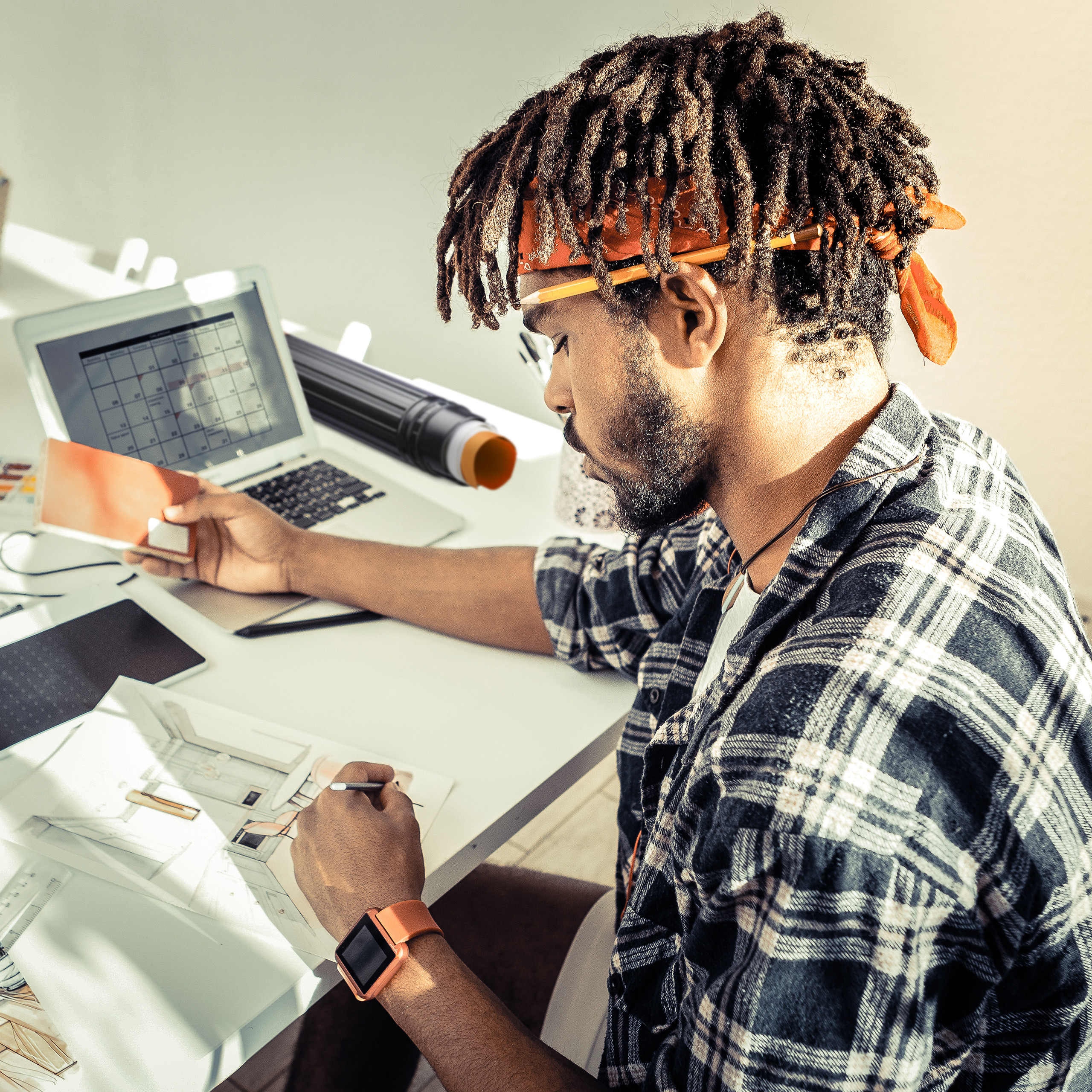 Ein junger Mann mit Dreadlocks und kariertem Hemd arbeitet an einem Schreibtisch und zeichnet Skizzen auf Papier.