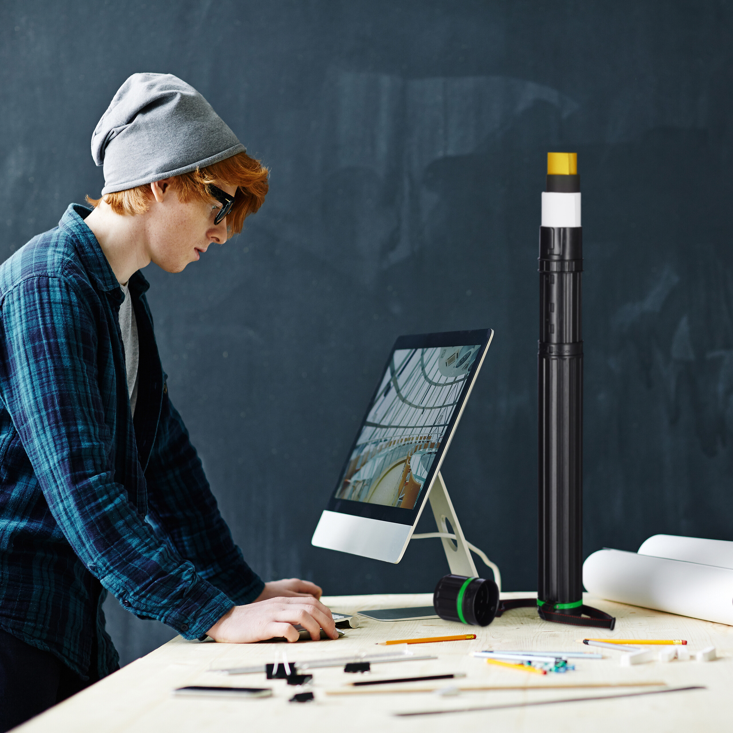 Ein rothaariger Mann mit grauer Mütze und Brille arbeitet an einem Computer auf einem Holztisch mit Zeichnungen.