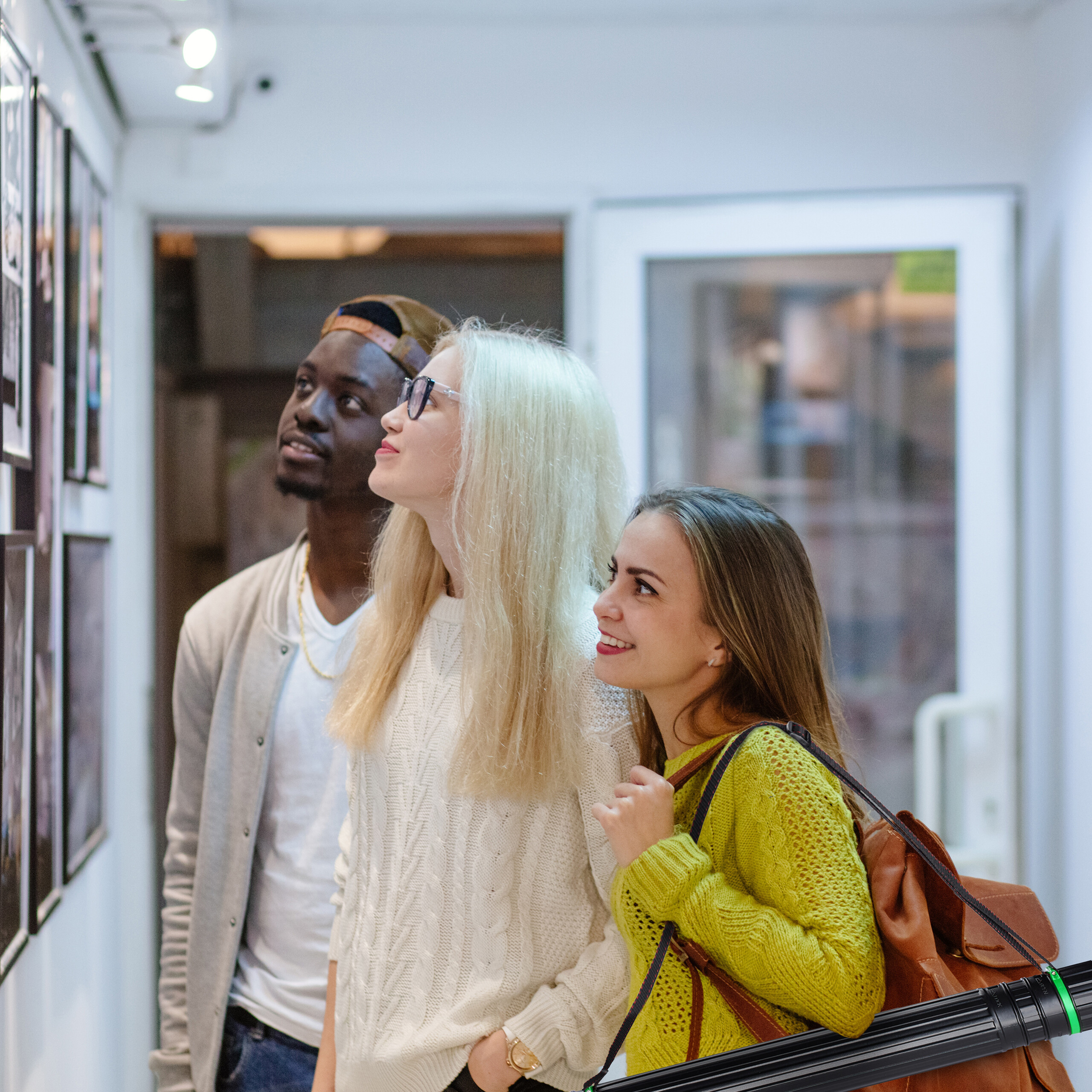 Drei diverse Freunde betrachten kunstvoll gerahmte Fotografien an einer weißen Wand in einer hellen Galerie.