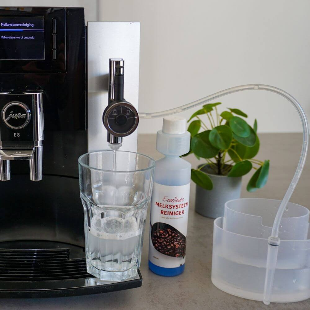 A Jura coffee machine cleans its milk system into a glass, with cleaner bottle and container nearby.