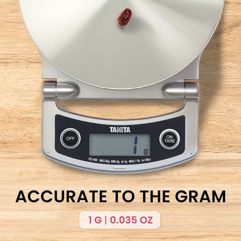 A single raisin sits on a silver kitchen scale on a wooden table, with the display reading 1g.