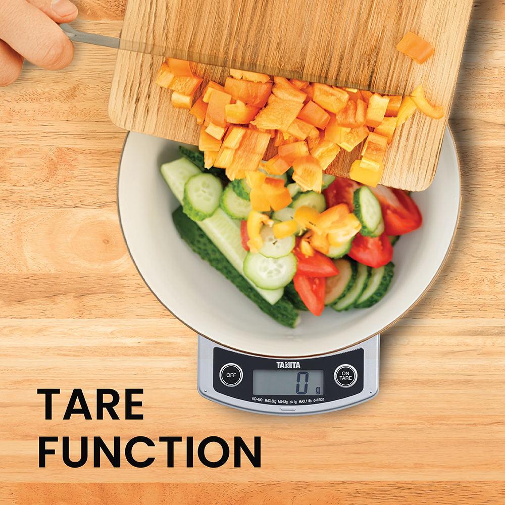 Chopped bell peppers sliding from a wooden board into a bowl of vegetables on a digital kitchen scale.