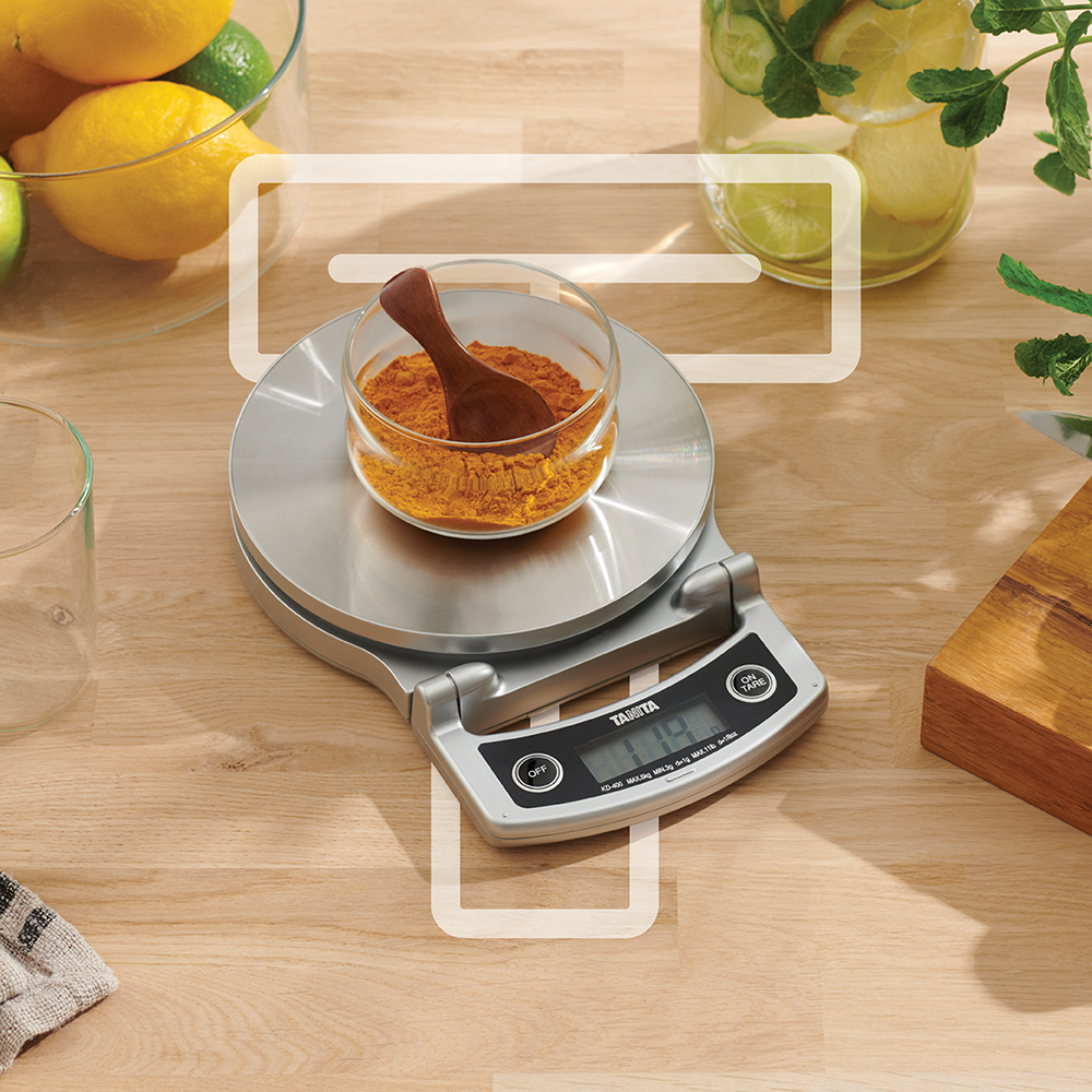 A digital kitchen scale with a bowl of turmeric and a wooden spoon on a wooden countertop.