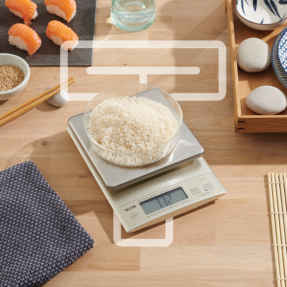 A metal kitchen scale on a wooden surface with a glass bowl of rice and sushi nearby.