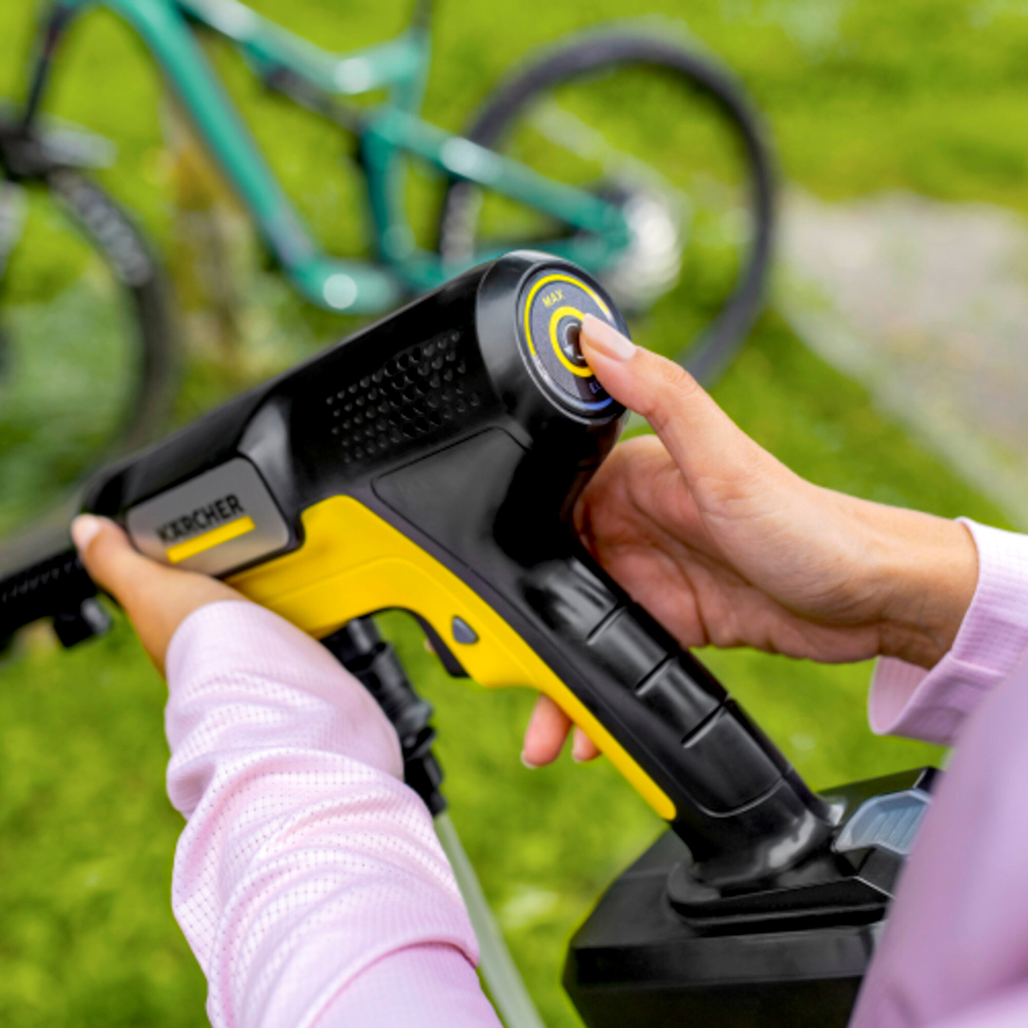 Eine Person in einem rosa Hemd hält einen Karcher-Druckreiniger, im Hintergrund ist ein Fahrrad zu sehen.