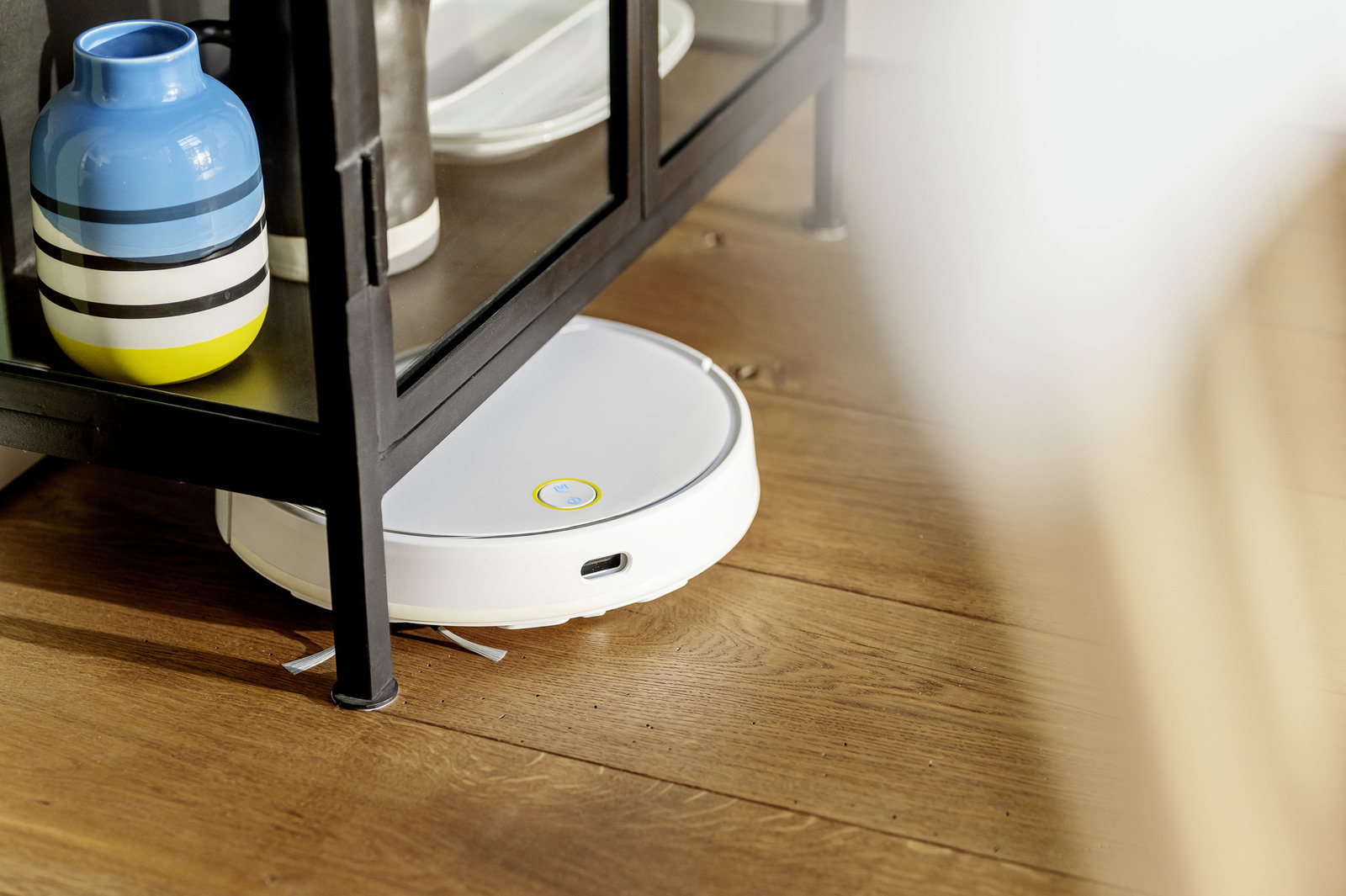 Un robot aspirador blanco limpia el suelo de madera bajo un mueble de metal negro con un jarrón colorido.