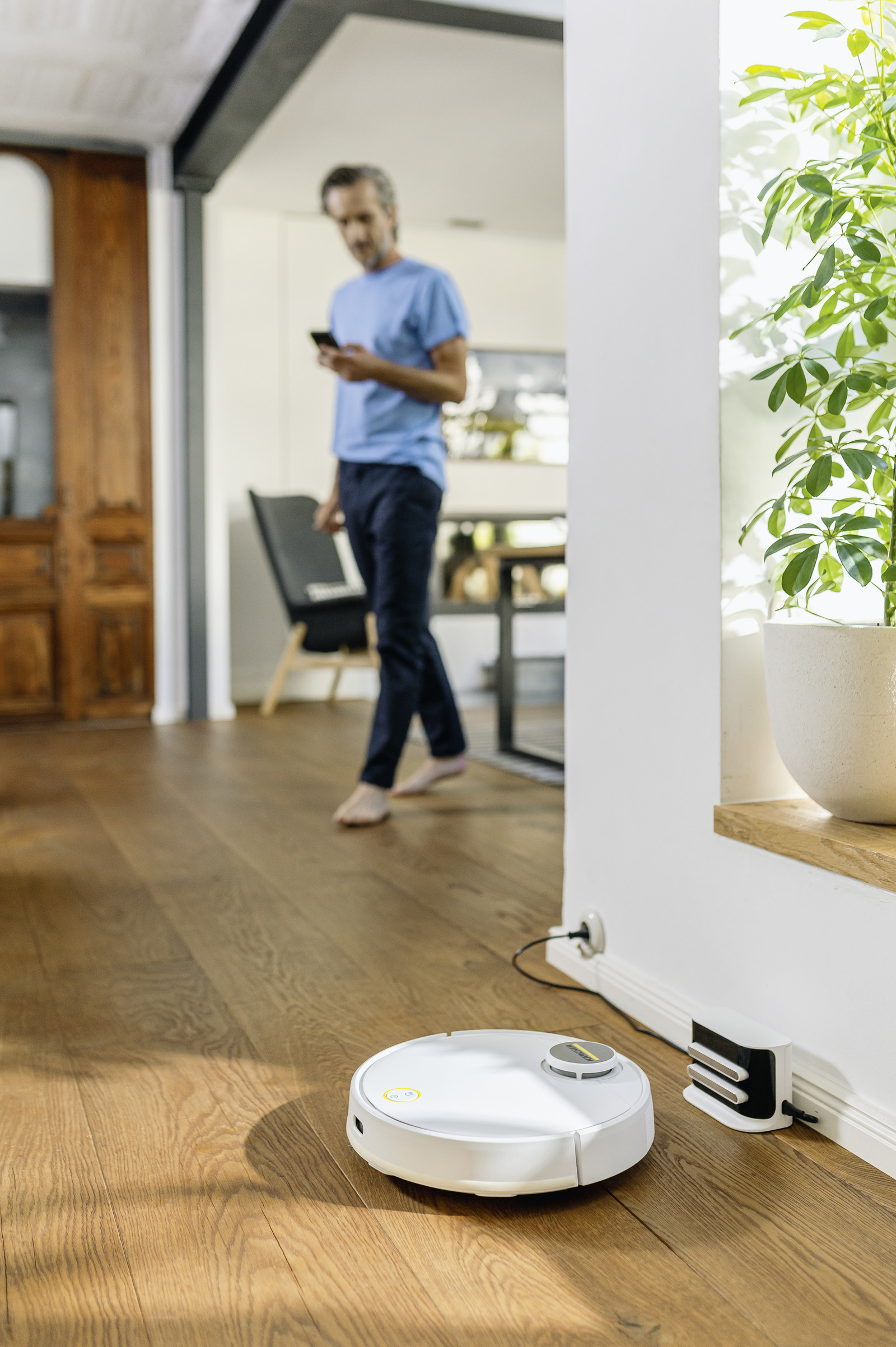 Un robot aspirador blanco se carga en un suelo de madera mientras un hombre camina al fondo.