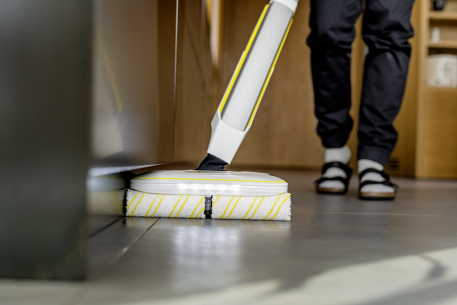 Eine Person nutzt einen weißen und gelben Bodenwischer auf grauen Fliesen in einem Raum.