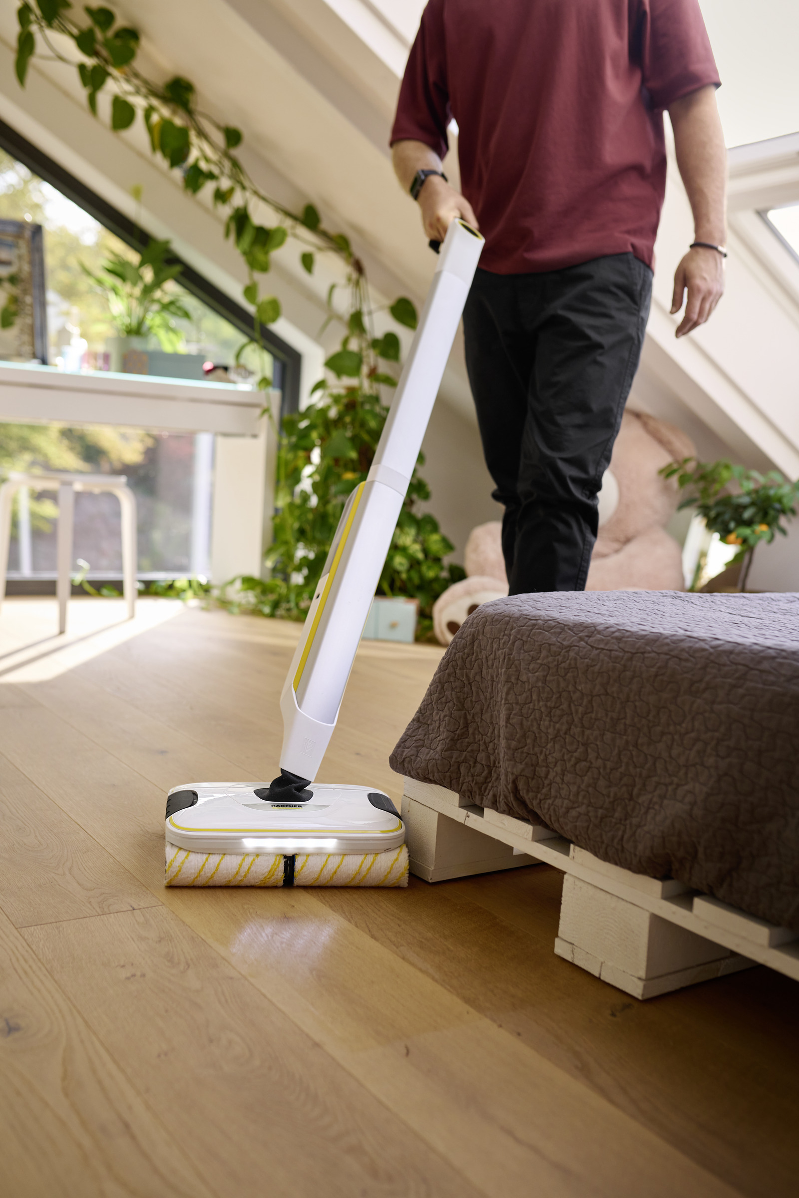 Ein Mann benutzt einen weiß-gelben Bodenreiniger auf Holzboden neben einem Bett.