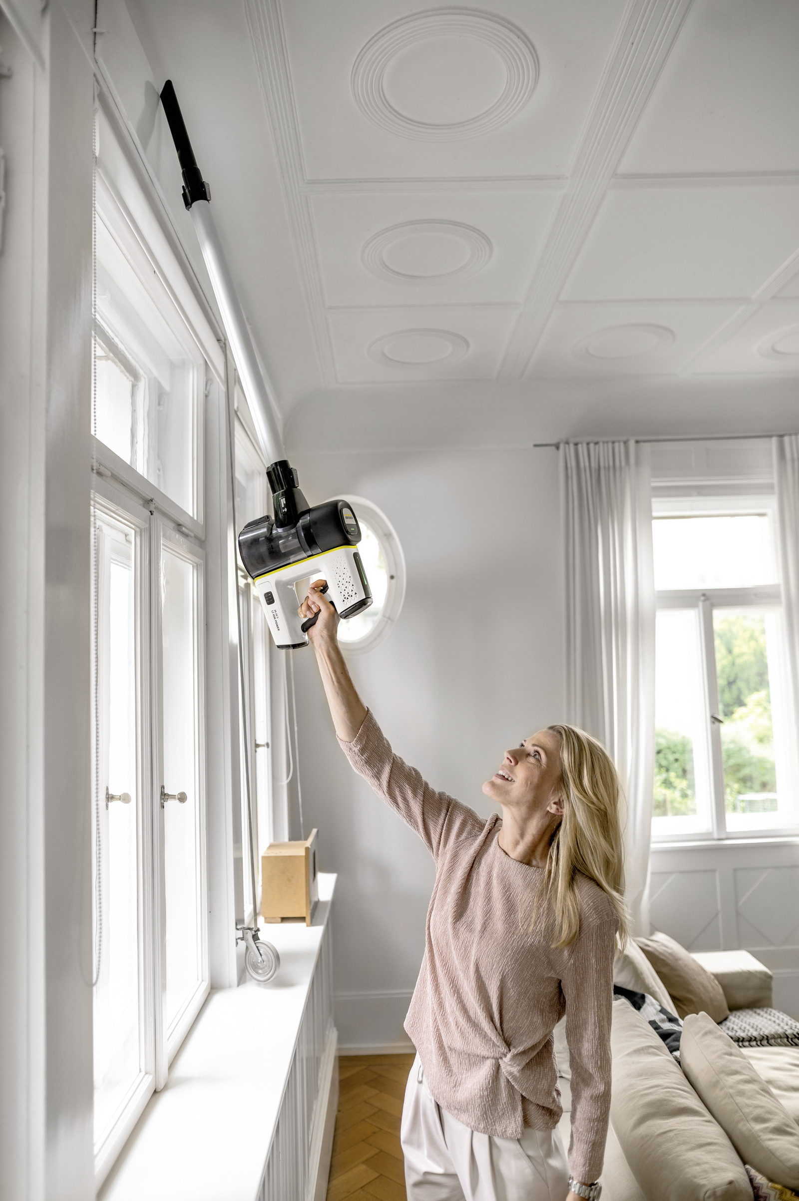 Una mujer rubia con un suéter rosa usa una aspiradora inalámbrica para limpiar el marco superior de la ventana en una habitación luminosa.